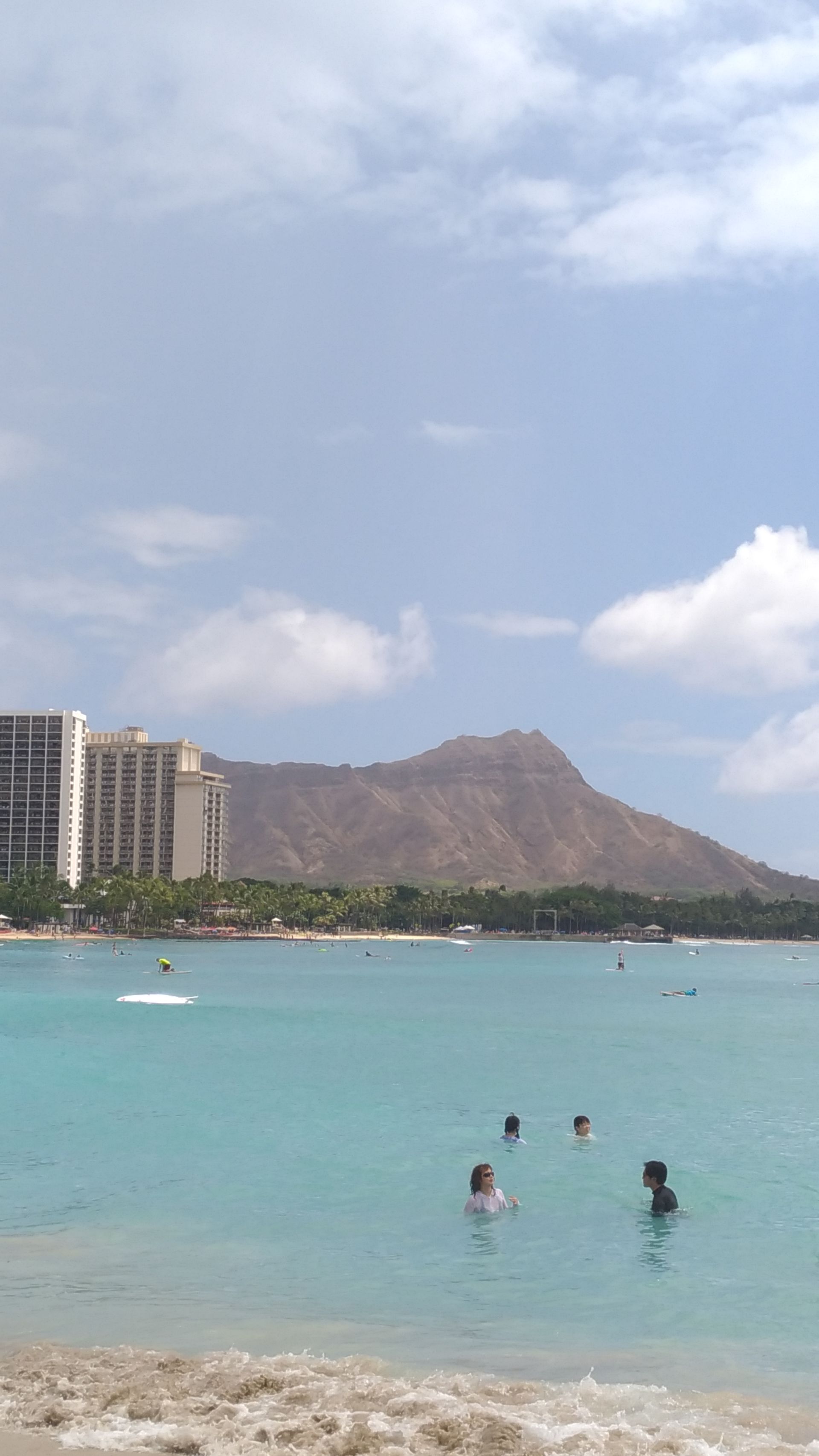 Ocean view with people swimming, buildings, and Diamond Head under a partly cloudy sky.