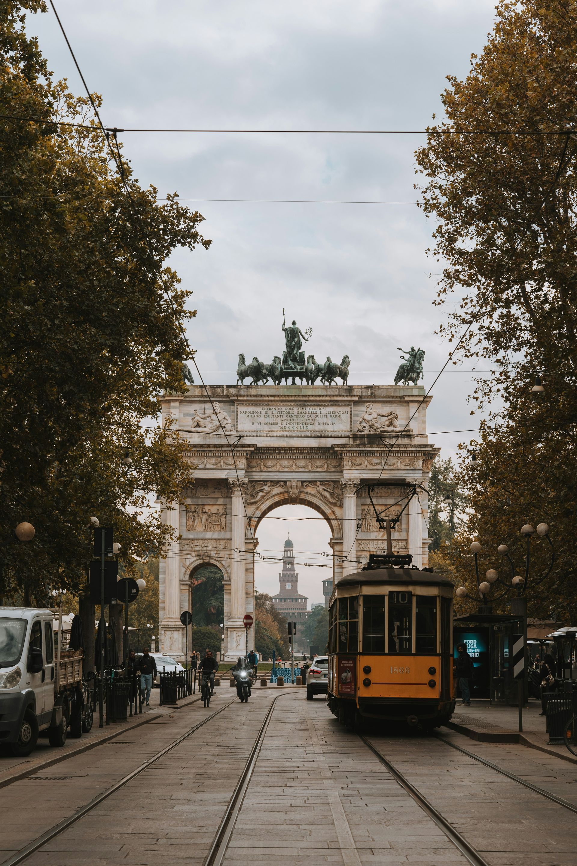 A trolley is driving down a street in Milan, in front of the large archway Arco della Pace.