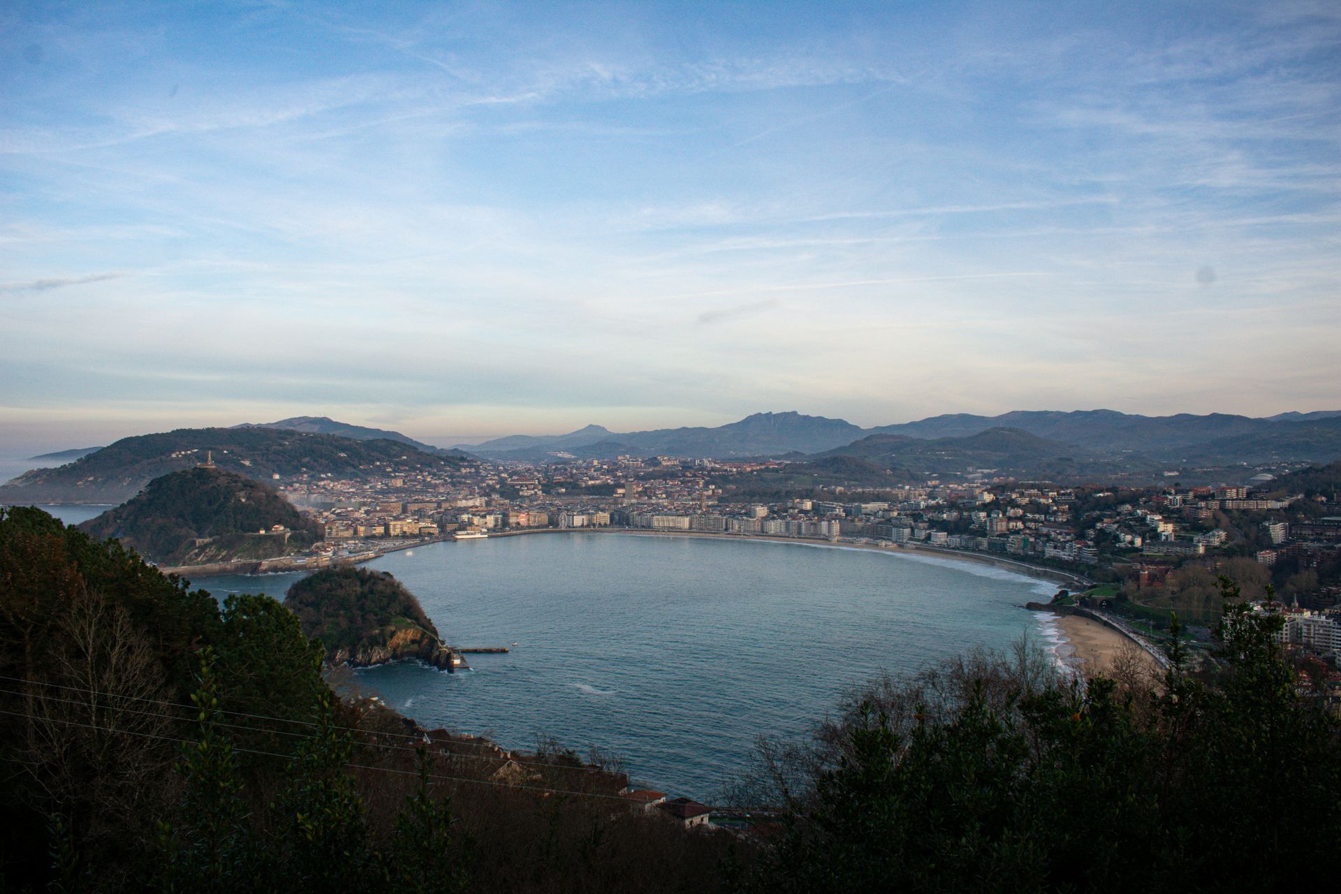 Panoramic view of San Sebastián, Spain, with a bay, beach, and city nestled between green hills and a blue sky.