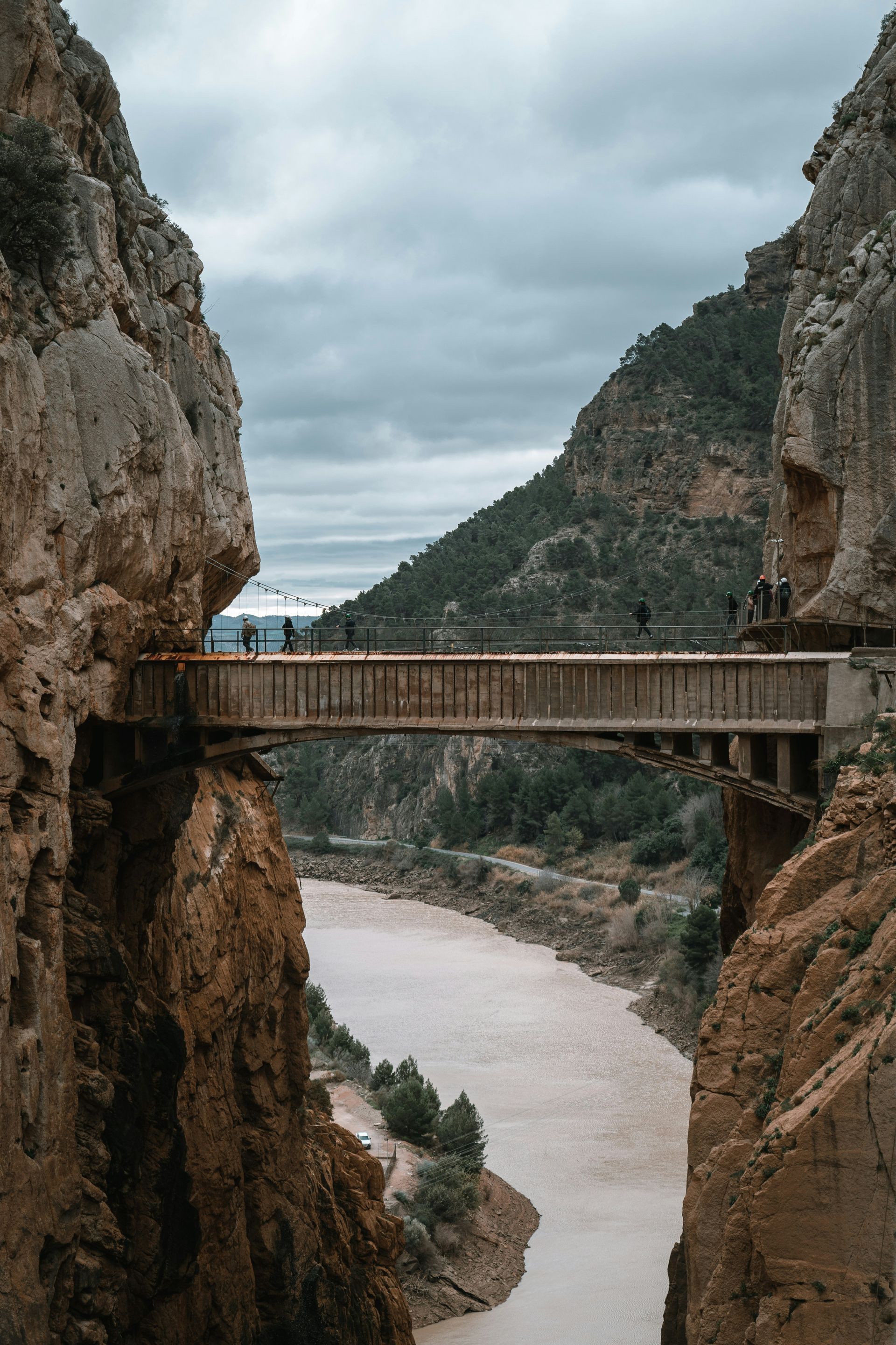 Bridge across a canyon with people walking. Brown water flows below, cliffs on both sides in Málaga, Spain.