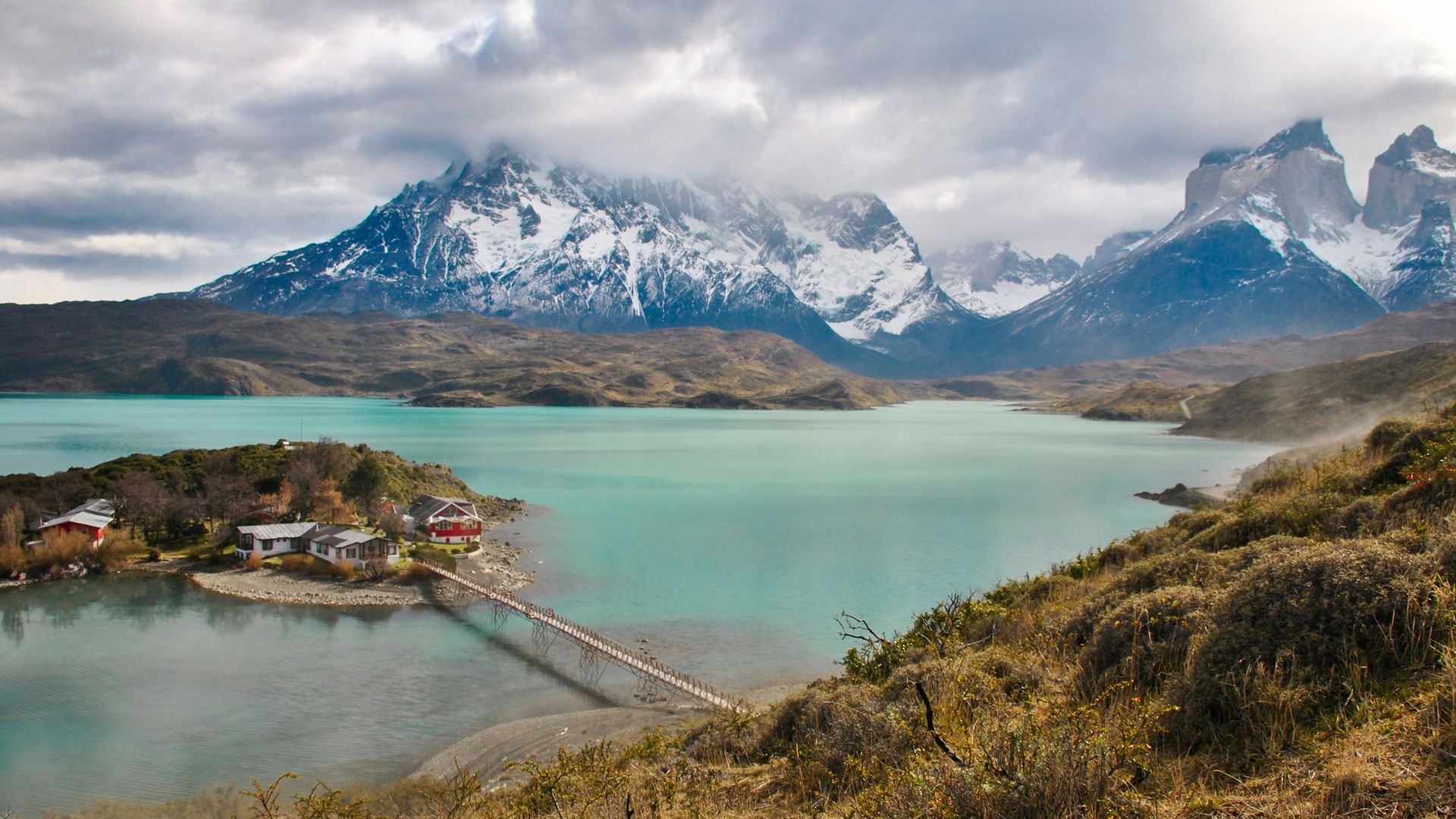 Turquoise lake with bridge, small buildings, and snow-capped mountains under a cloudy sky in Torres del Paine, Chile.