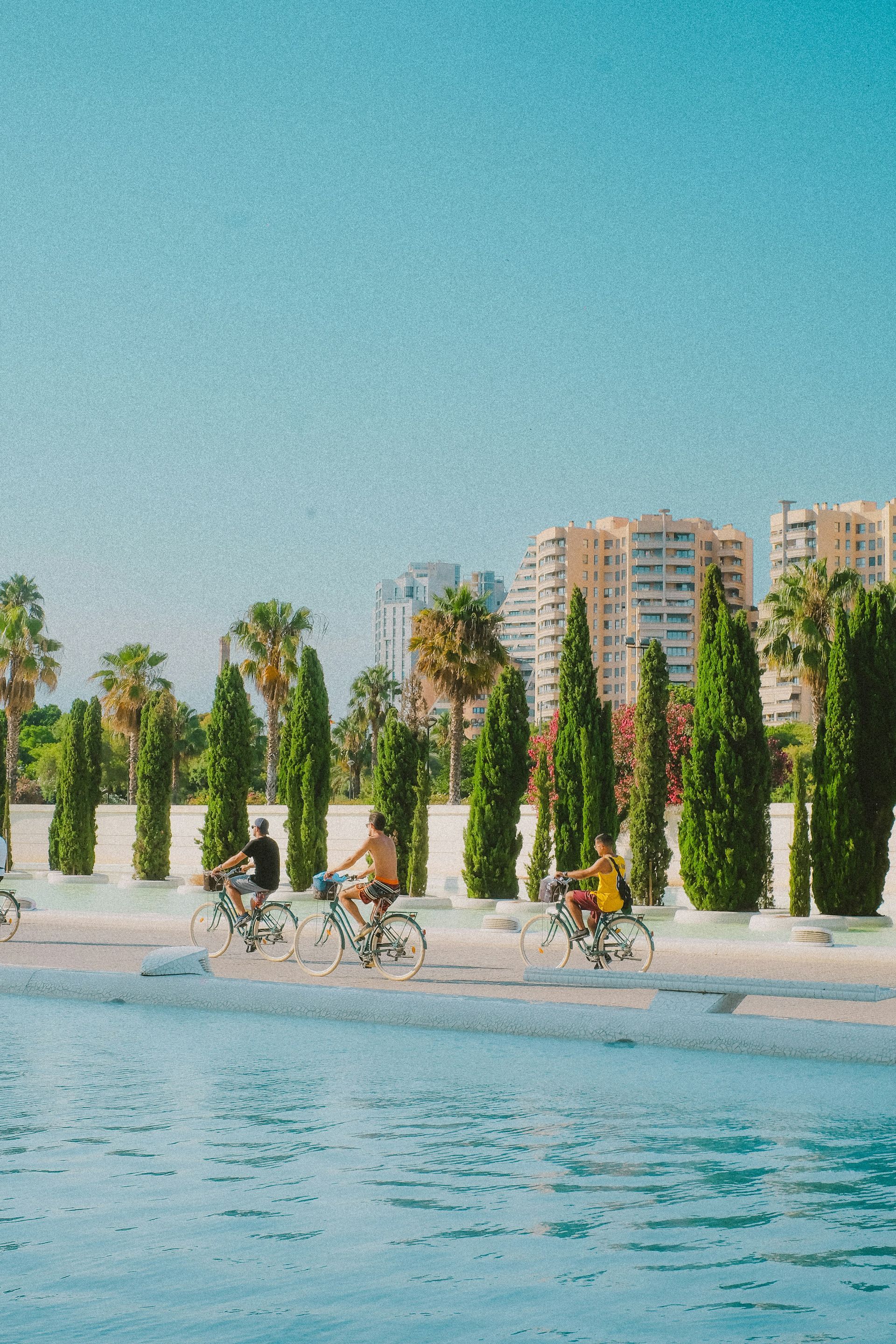 Three cyclists riding on a path next to water and trees, with buildings in the background in Valencia, Spain.