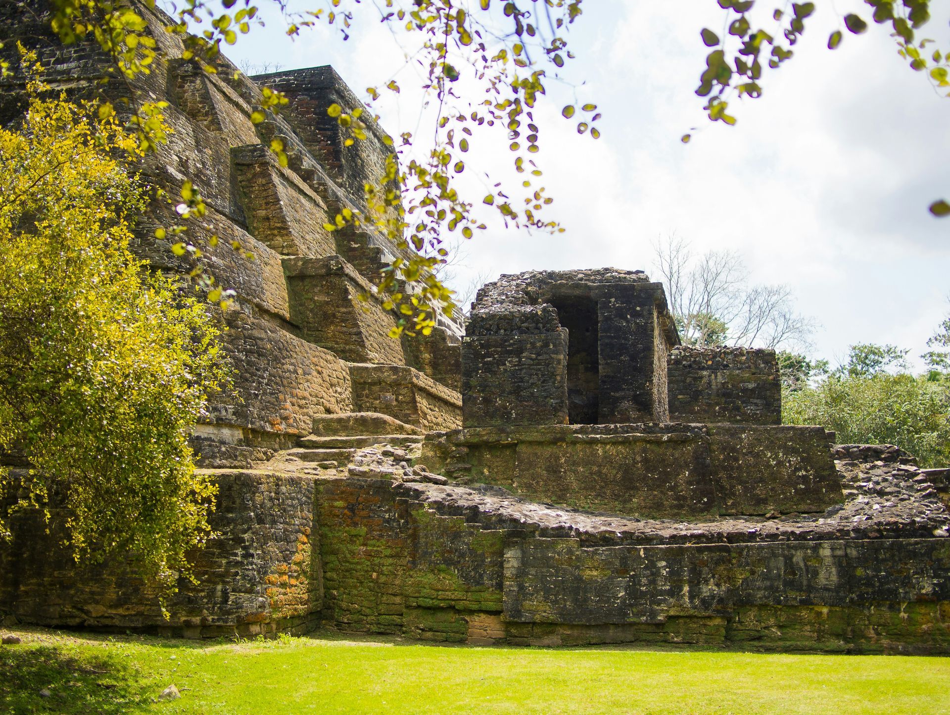 Ancient Mayan ruins of Xunantunich, Belize. A weathered stone structure sits amidst green grass and trees, with a cloudy sky.