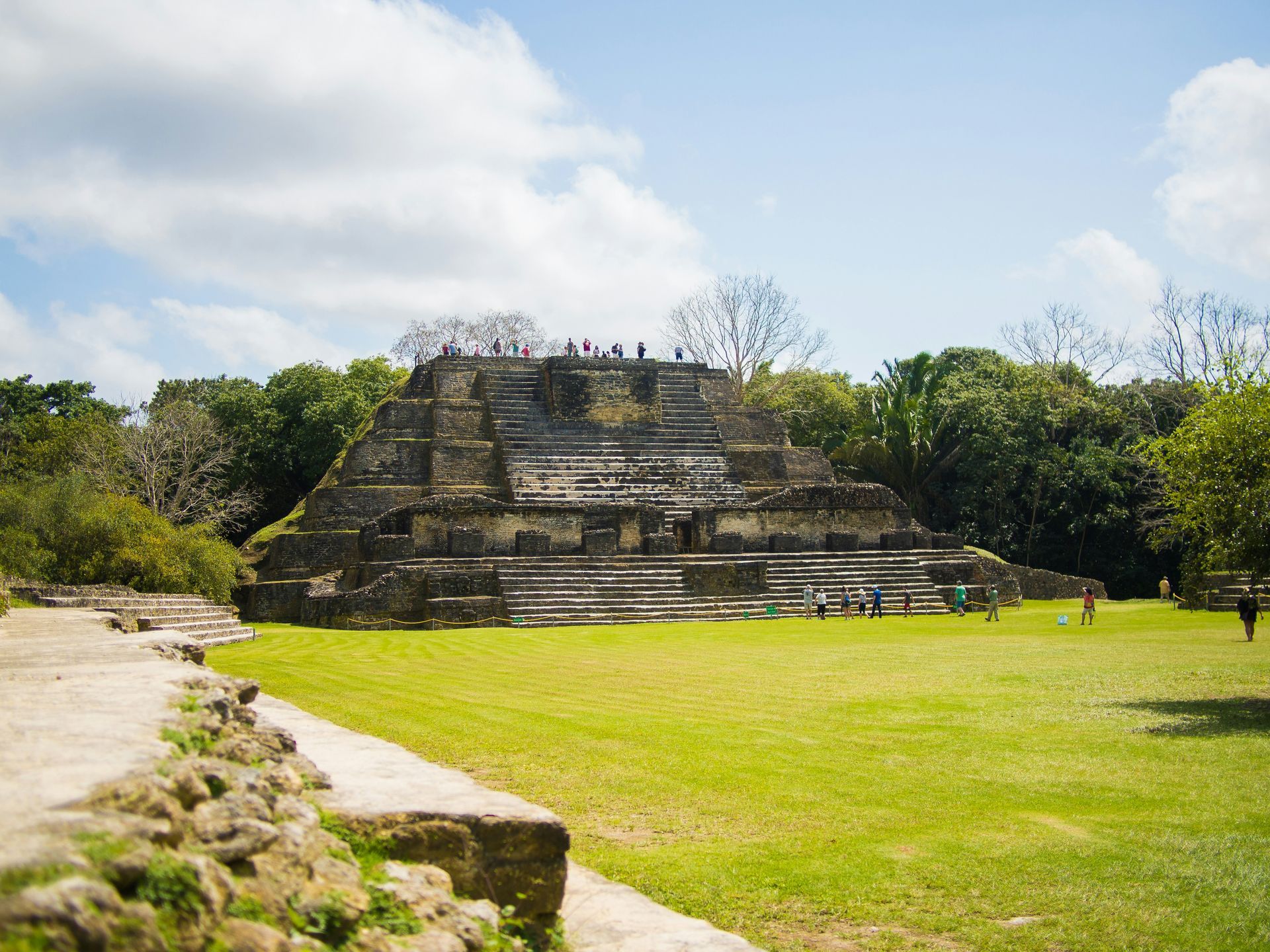 Ancient Mayan temple with tiered structure on a grassy field, people exploring, surrounded by trees in Belize.