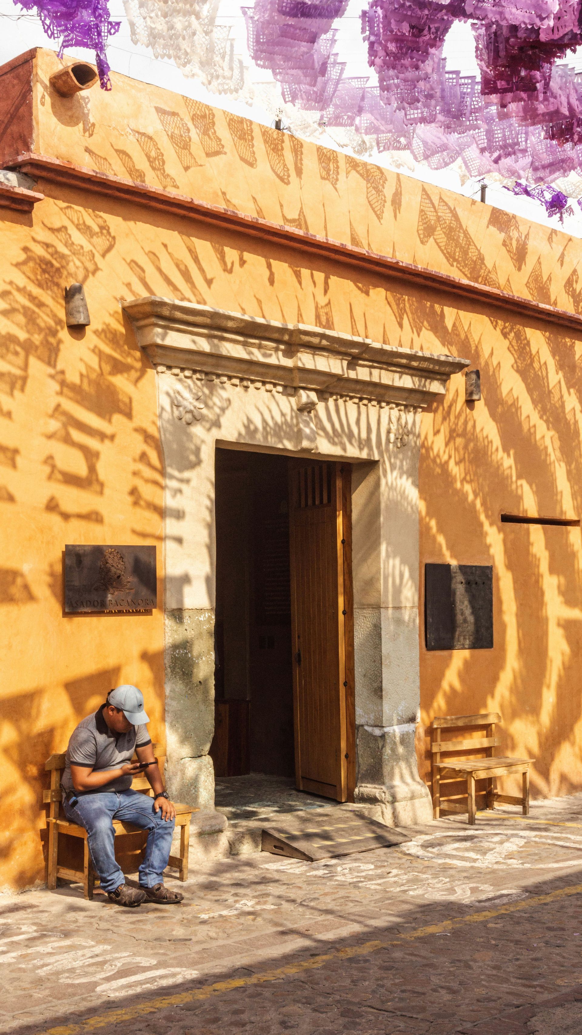 Man sits on a wooden chair in front of a yellow building with an open doorway in Oaxaca, Mexico.