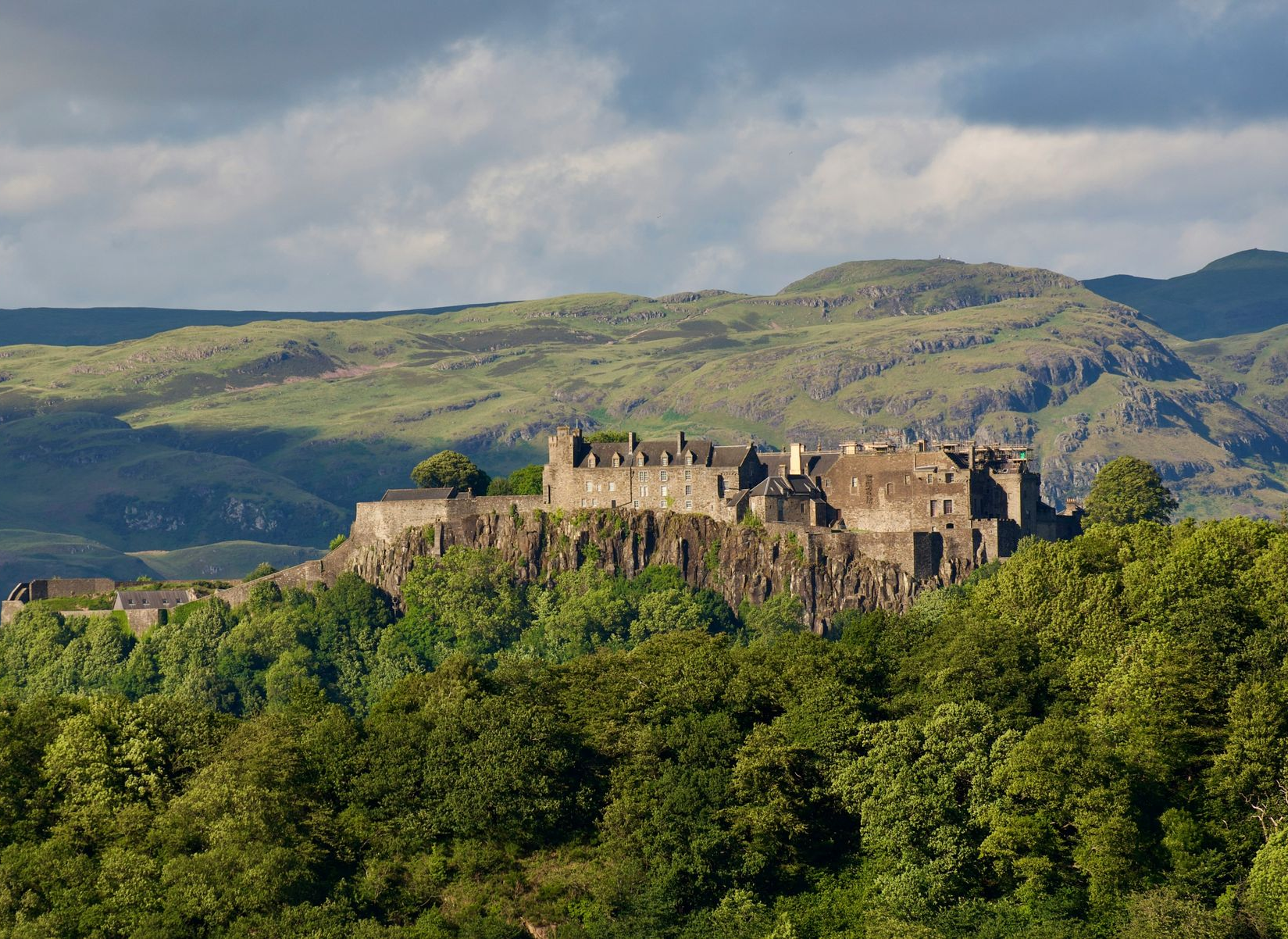 The Stirling Castle ruins atop a green, tree-covered hill with mountains in the distance under a cloudy sky in Scotland.
