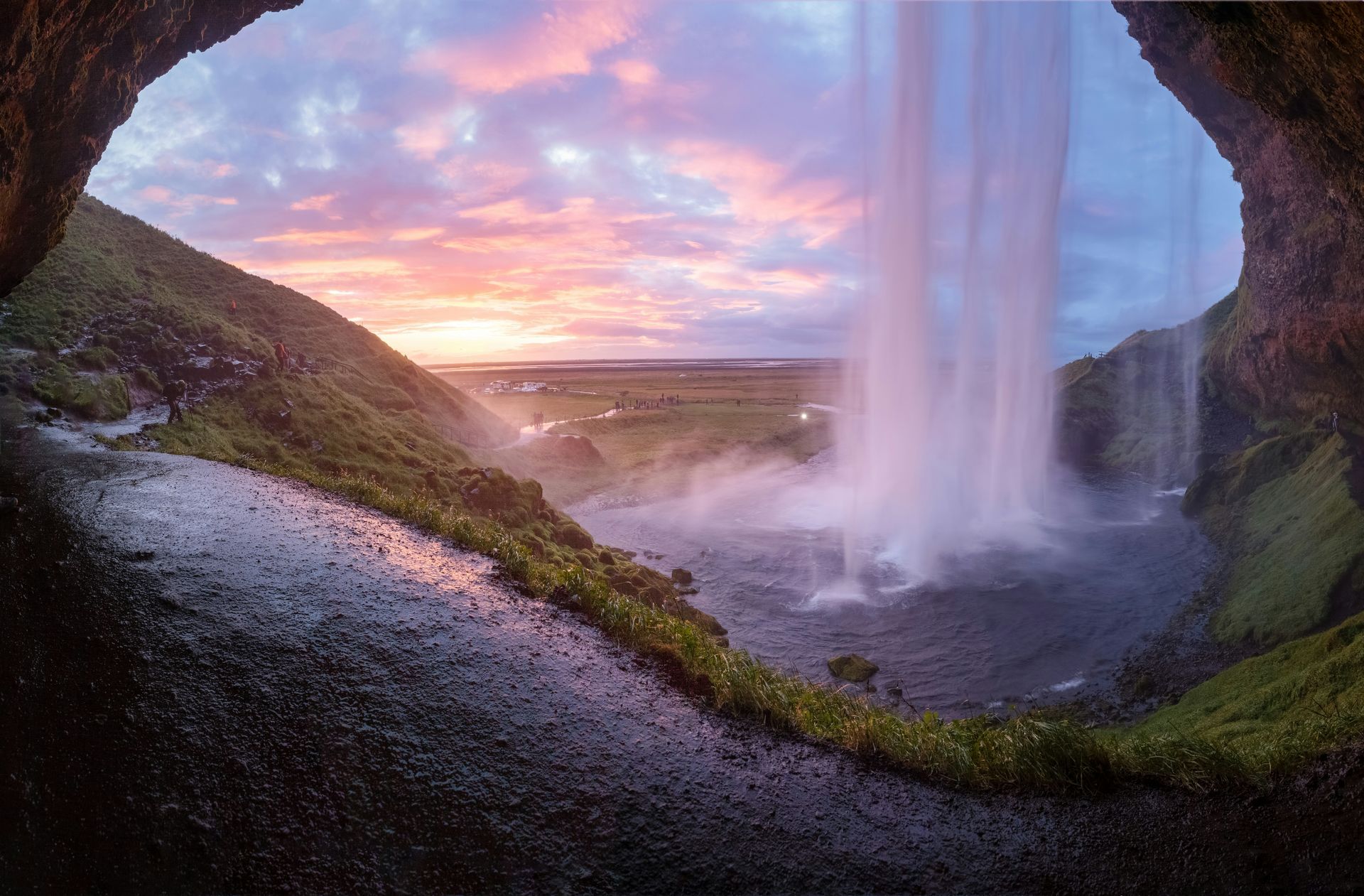 The Seljalandsfoss Waterfall at sunset, viewed from a cave. Pink and orange sky, green grass and rocks in Iceland.