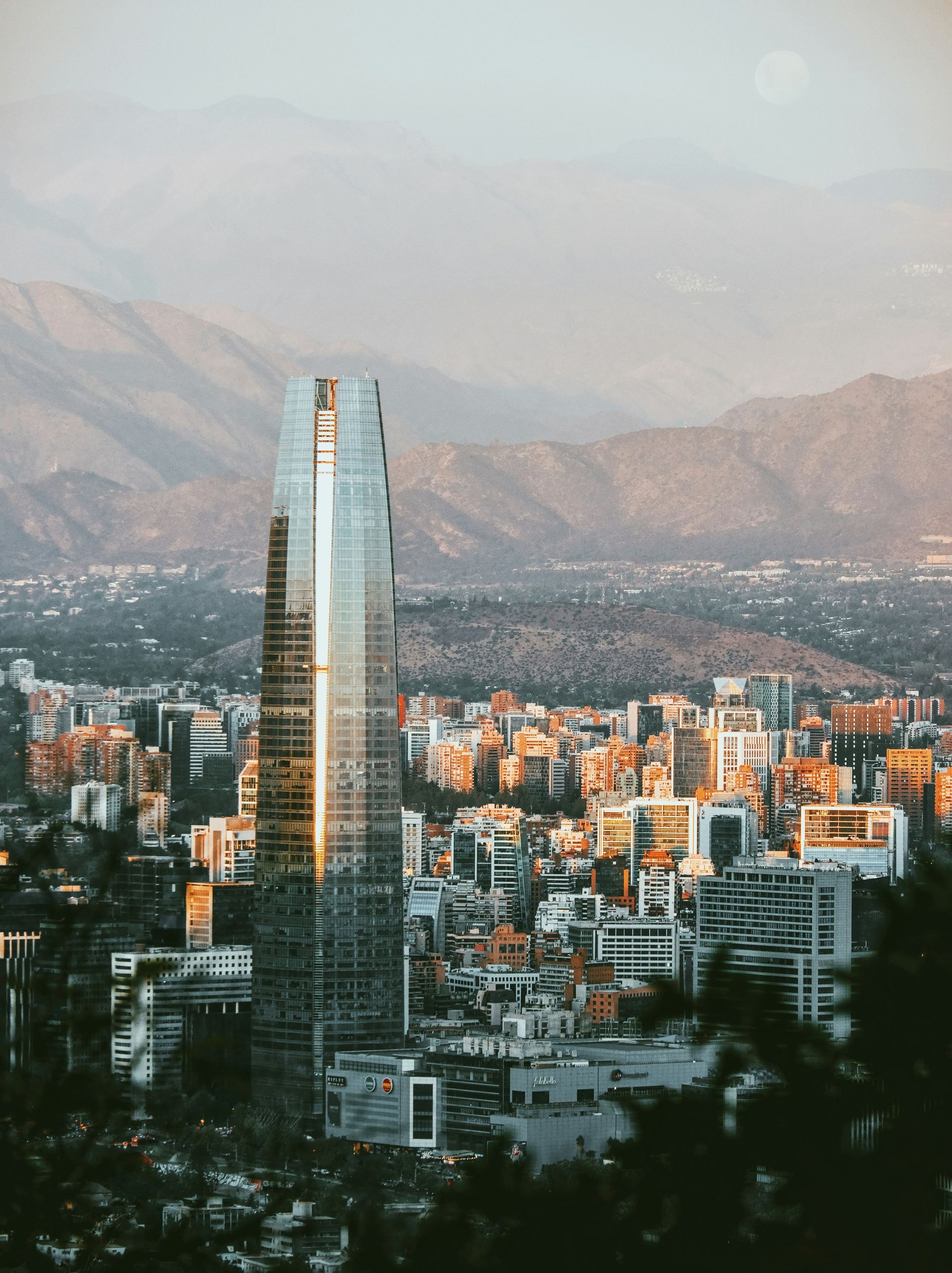 A glass skyscraper dominates a cityscape with mountains in the background. Golden light bathes the scene in Santiago, Chile.