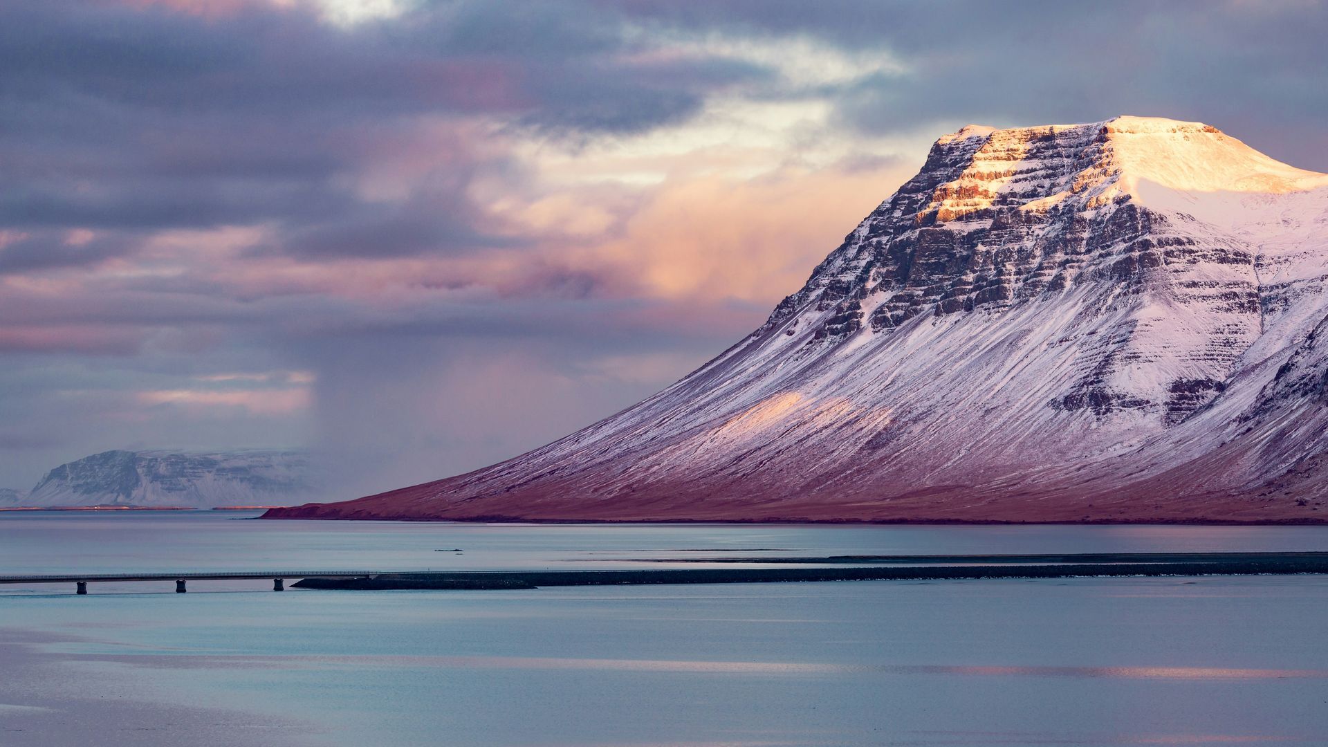 Snow-capped mountain with golden sunlight, reflected in water with a bridge, purple and pink sky in Snæfellsnes, Iceland.