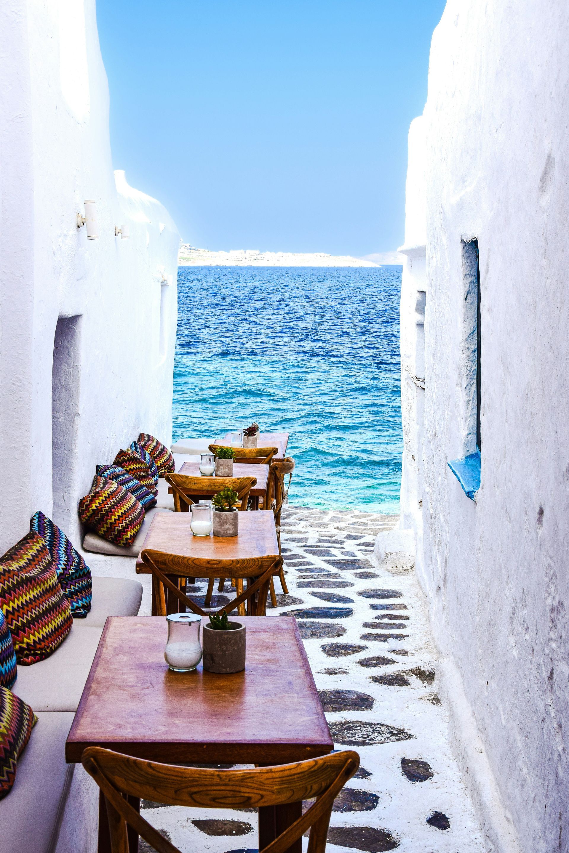 A narrow alleyway with tables and chairs leading to the ocean in Mykonos, Greece.