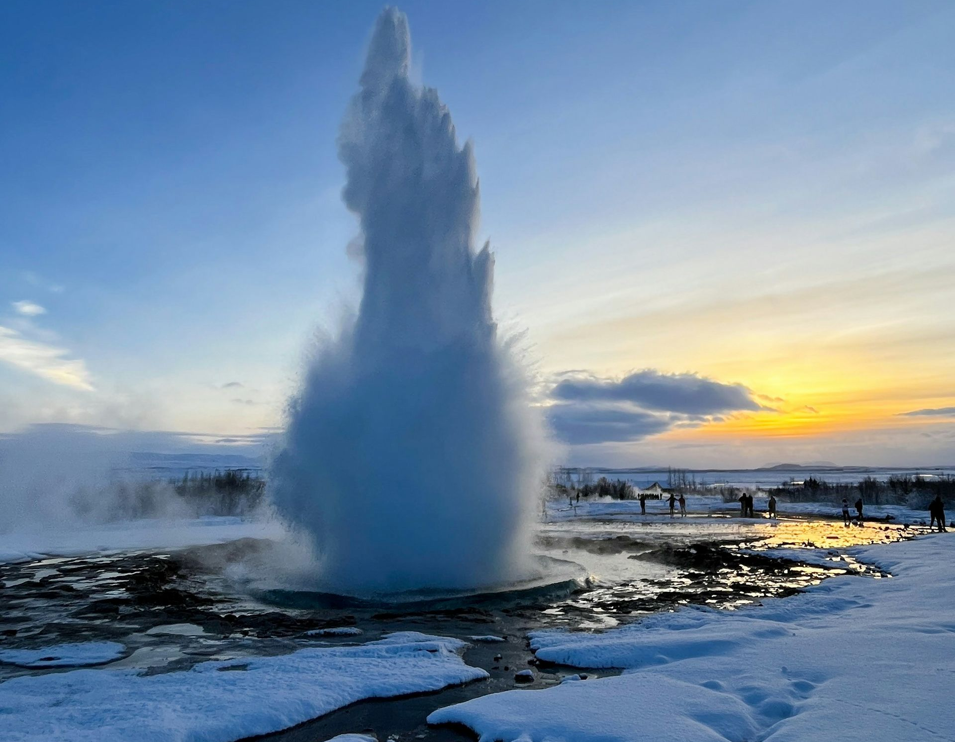 Geyser Strokkur erupts amidst snow-covered landscape under a blue and yellow sky in Iceland.