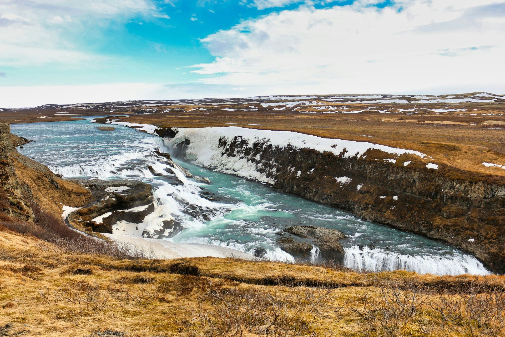 Gullfoss Falls cascading into a gorge; snow-covered banks, brown grass, blue sky in Iceland.
