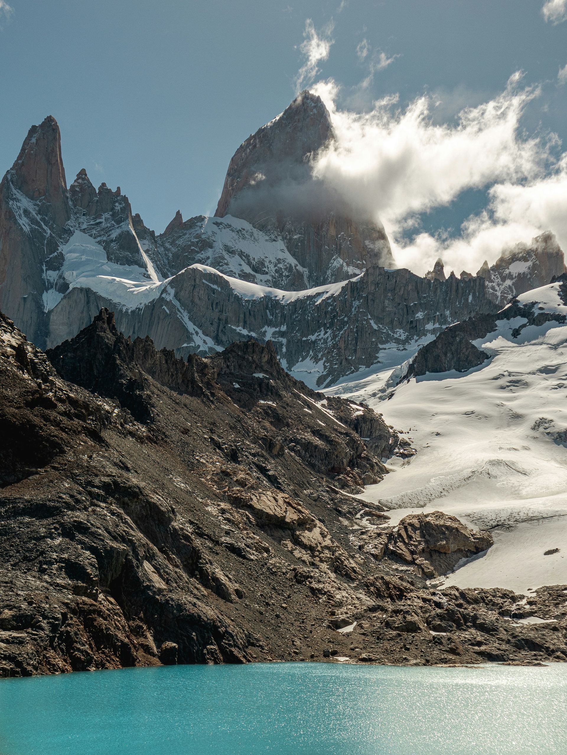 Snow-capped mountains rise above a turquoise lake under a cloudy sky in El Chaltén, Argentina.