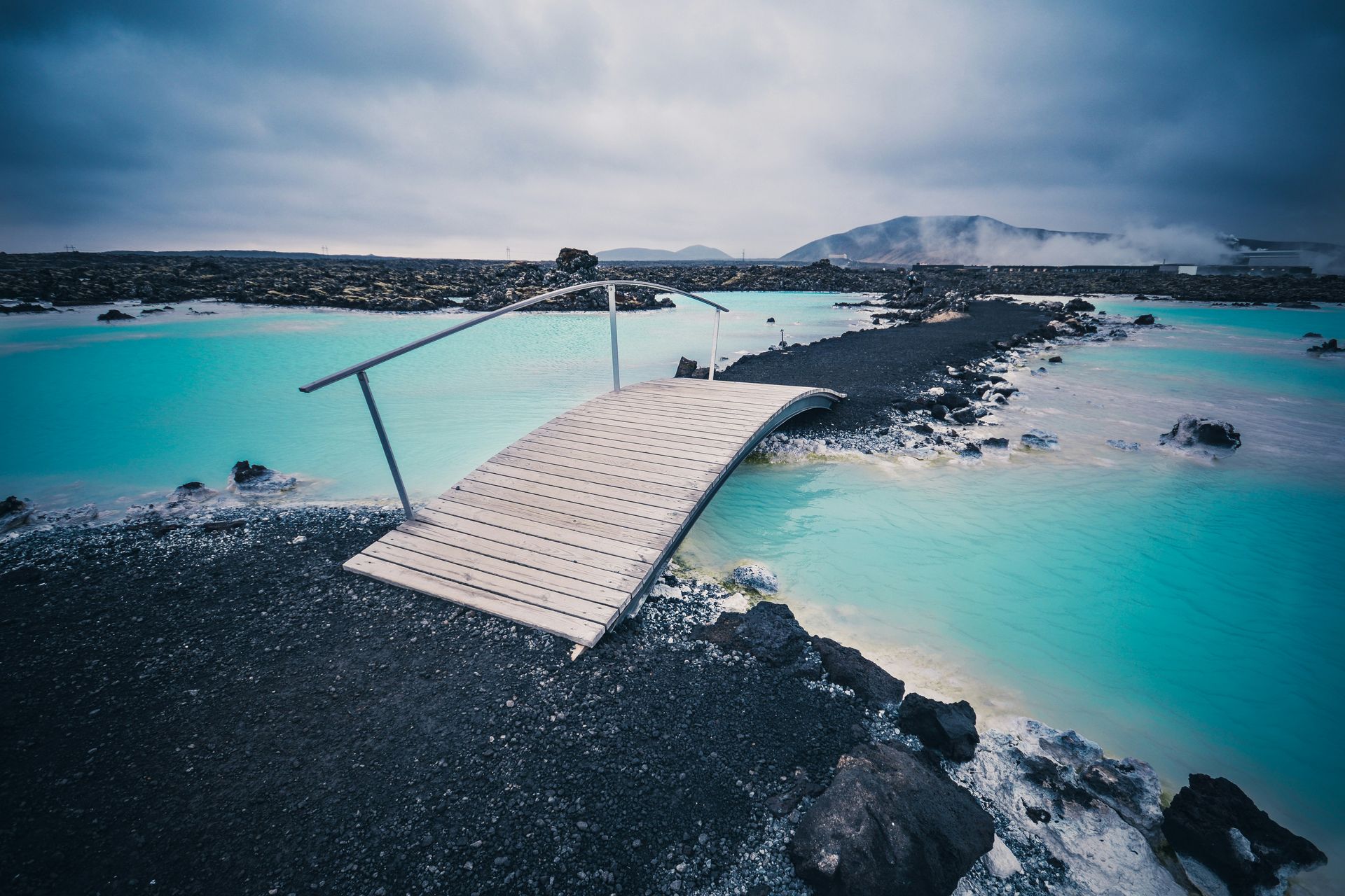 Wooden bridge over bright blue geothermal lagoon, dark rocks at Blue Lagoon, Iceland.