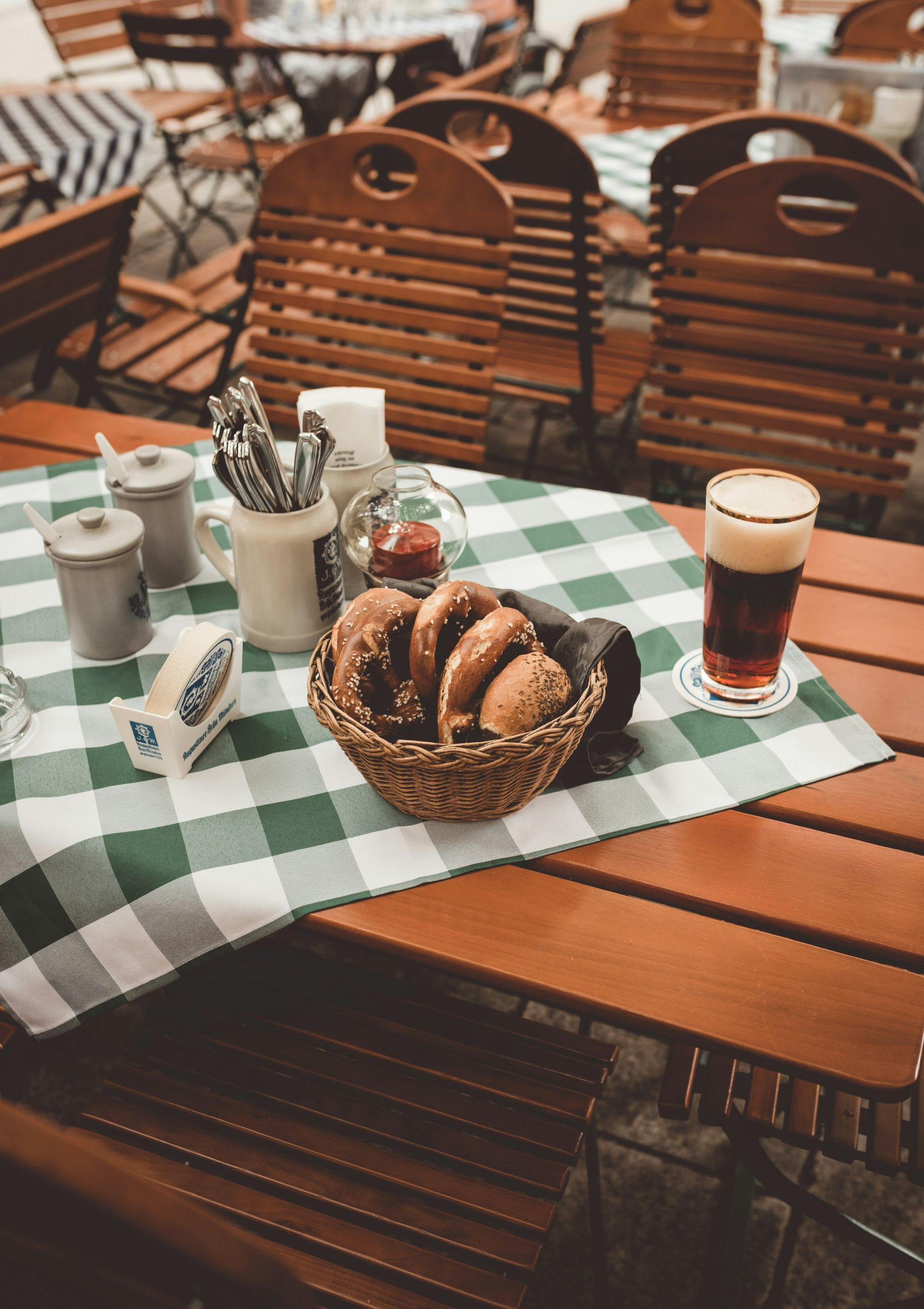 Outdoor cafe table with a basket of pretzels, beer, and condiments, and a green checkered tablecloth in Germany.