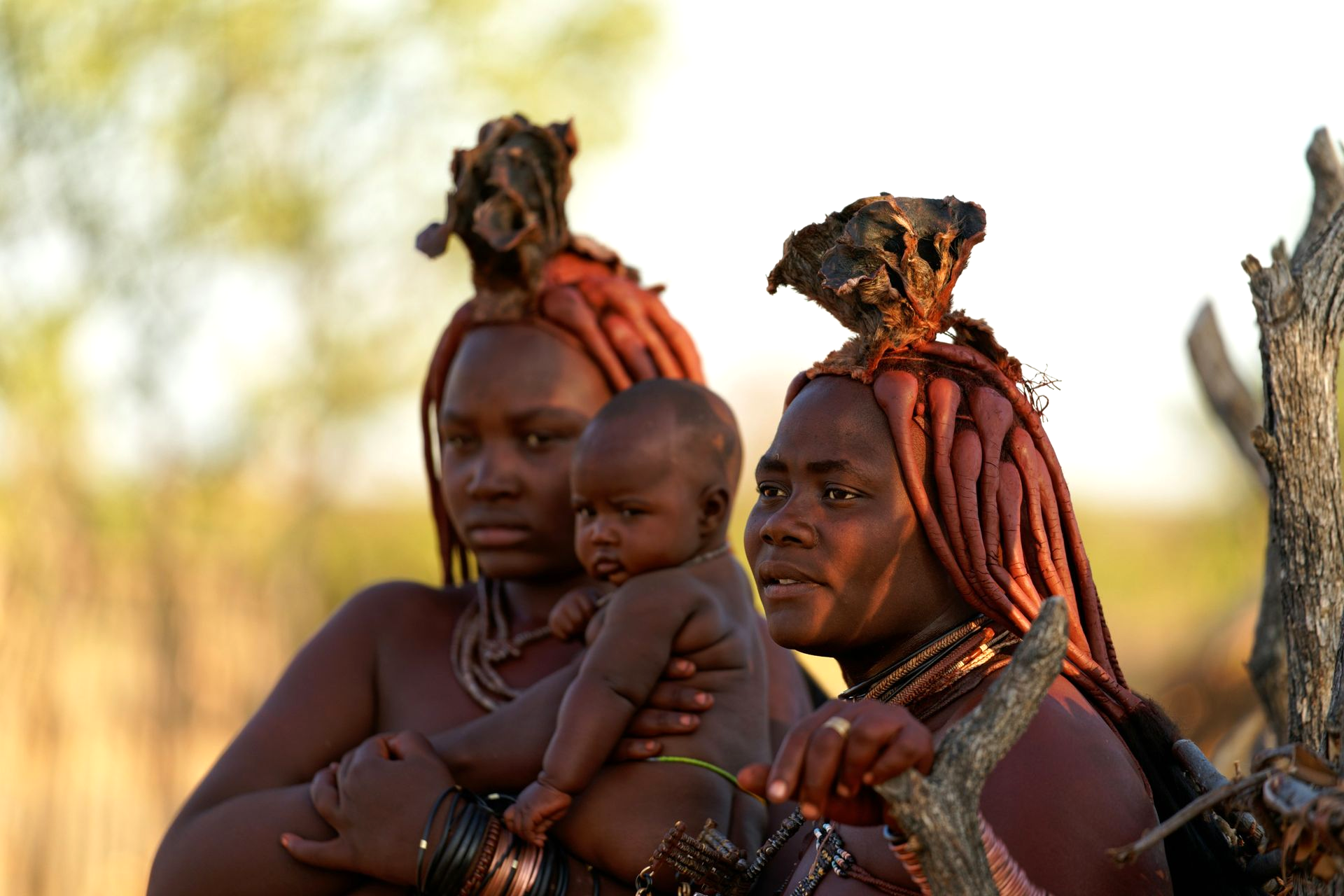 Two Himba women and a baby in Namibia, with red clay hairstyles and skin, holding the infant.