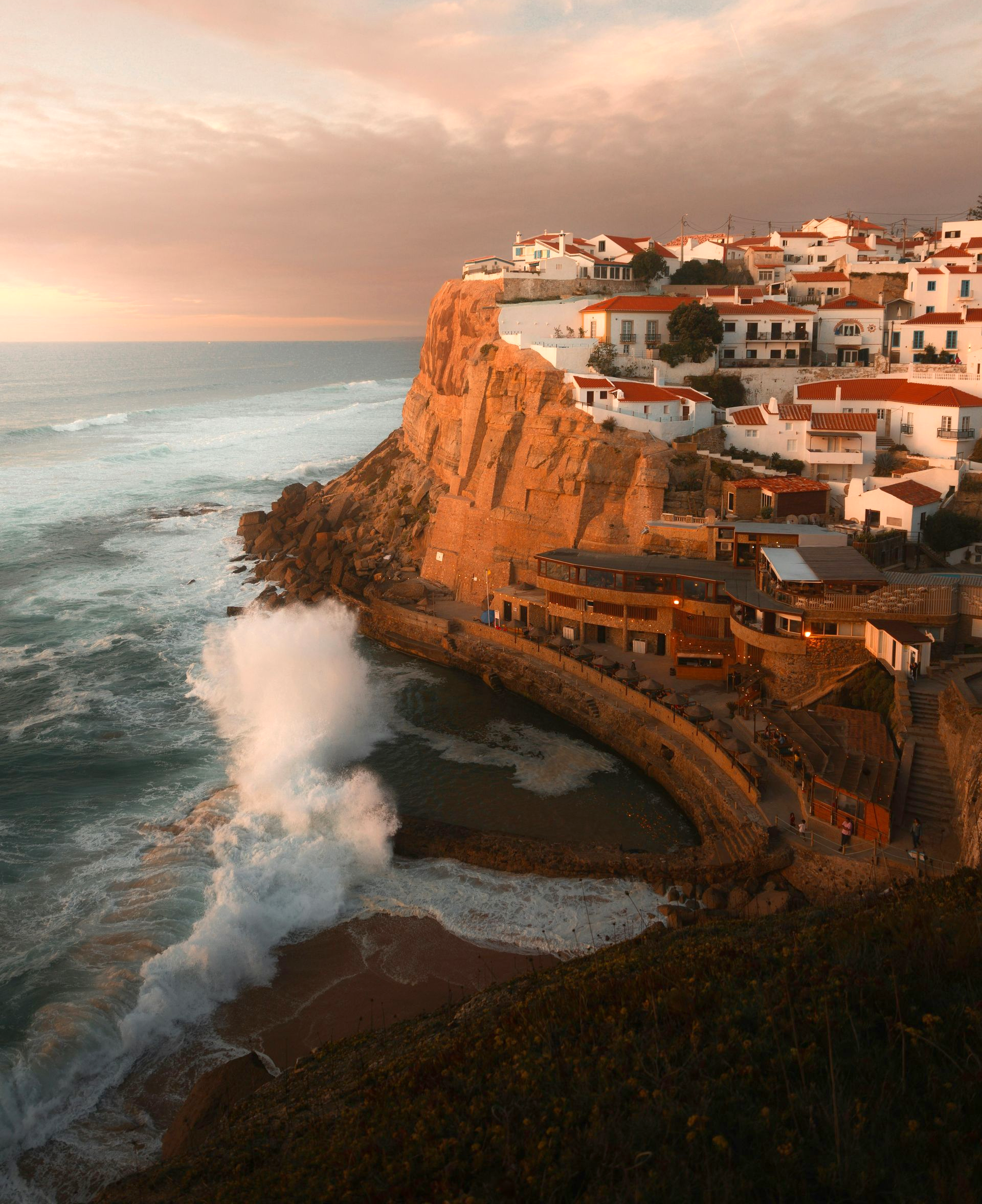 Village of Azenhas do Mar on a cliff at sunset; waves crash against the shore, houses with red roofs in Portugal.