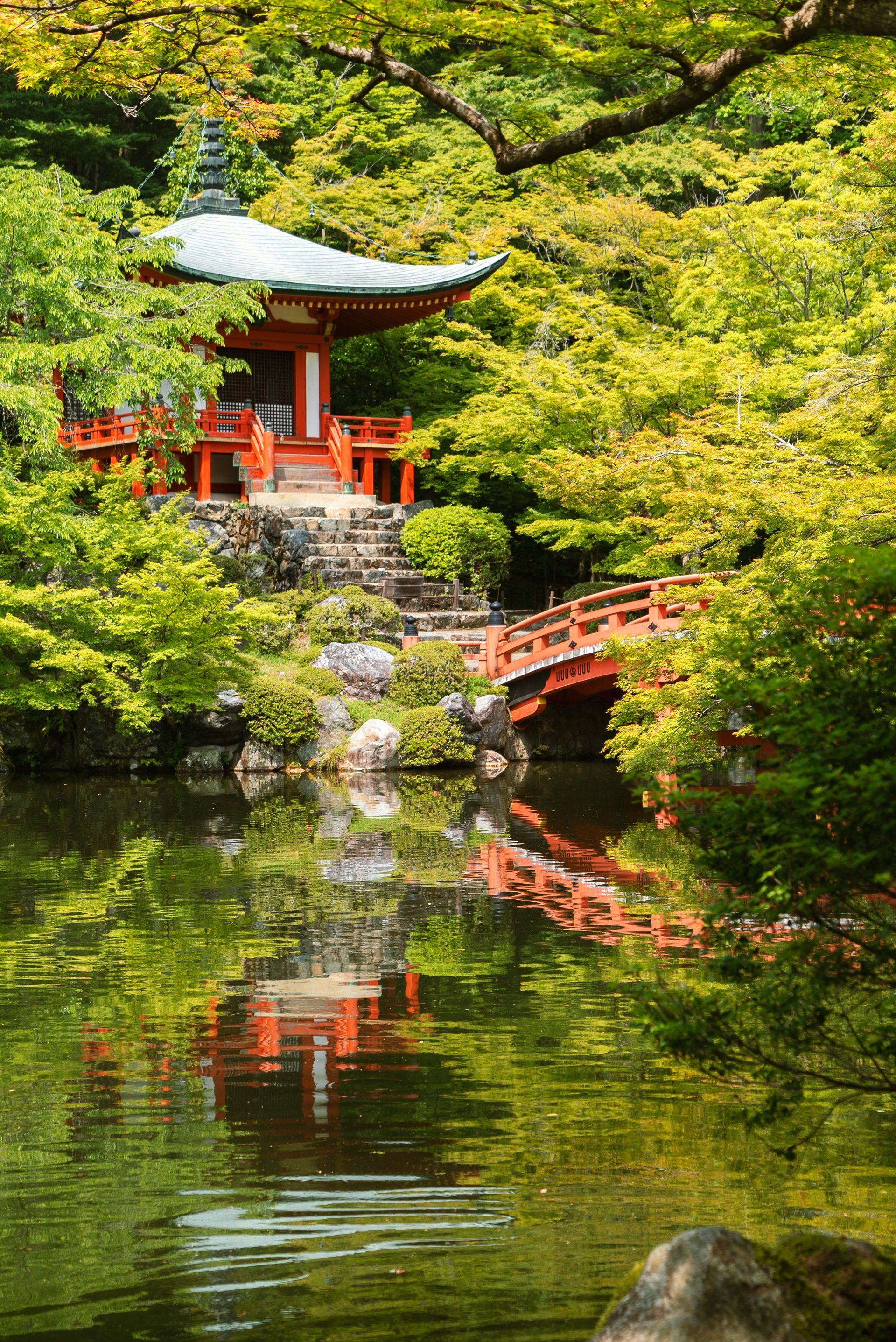 Red pagoda and bridge reflected in tranquil pond, surrounded by lush green foliage in Kyoto, Japan.