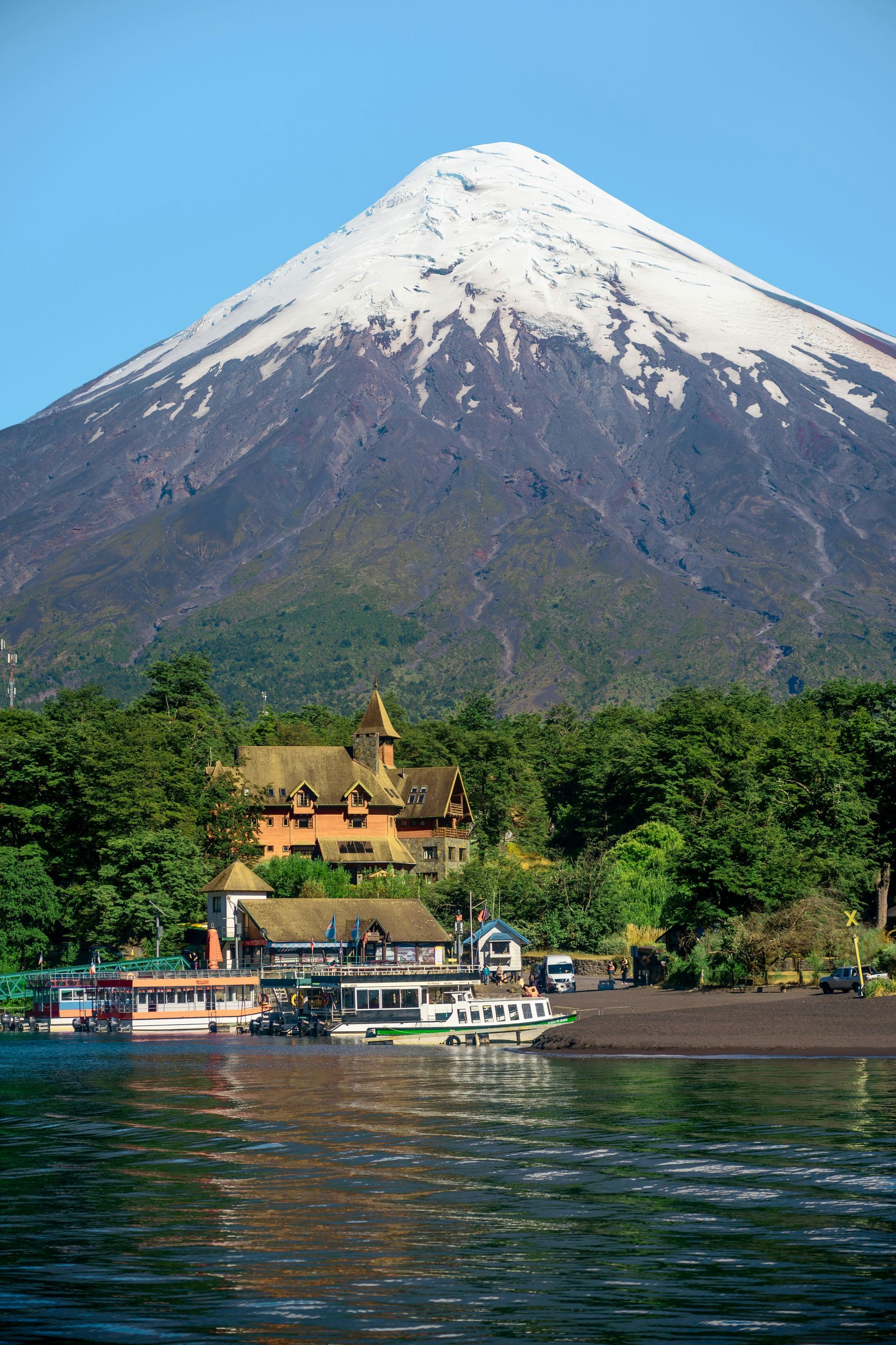 Snow-capped volcano towering above a lake with buildings and boats along the shore in Los Lagos, Chile.