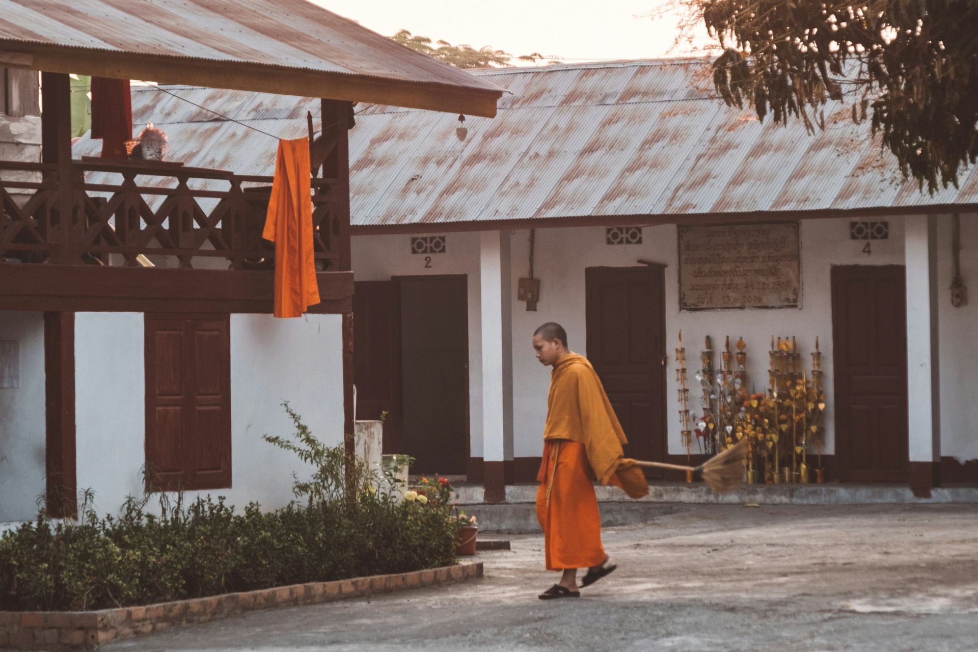 Monk in orange robes walks in a courtyard, near a building with corrugated metal roofing in Luang Prabang, Laos.
