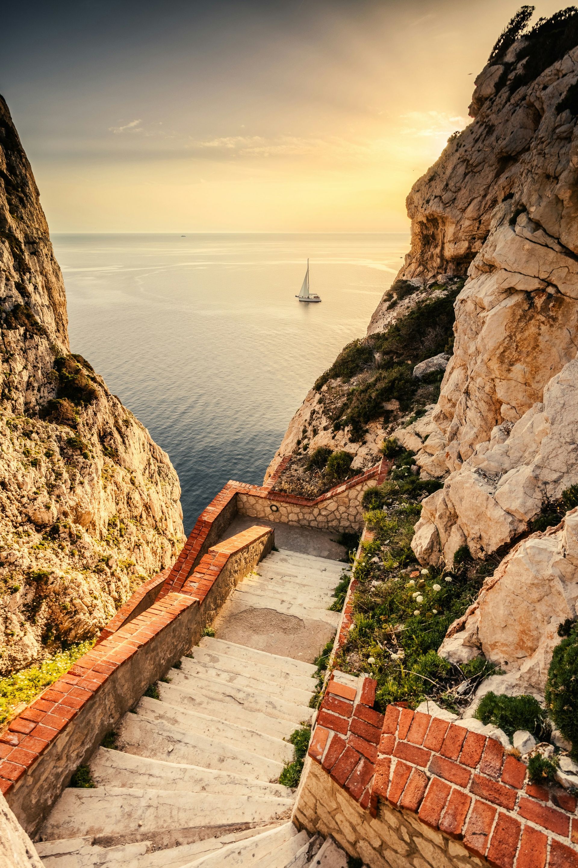 A staircase leading up to the ocean with a sailboat in the distance in Sardinia, Italy.