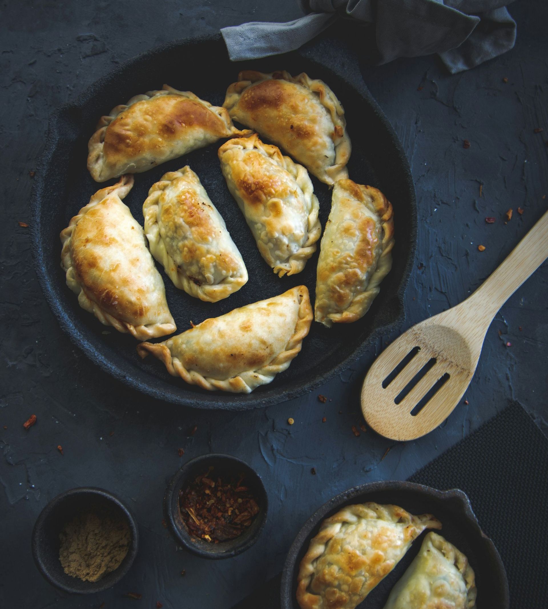 Golden-brown empanadas on a black plate with spice bowls, a wooden spoon, and a dark background in Argentina.