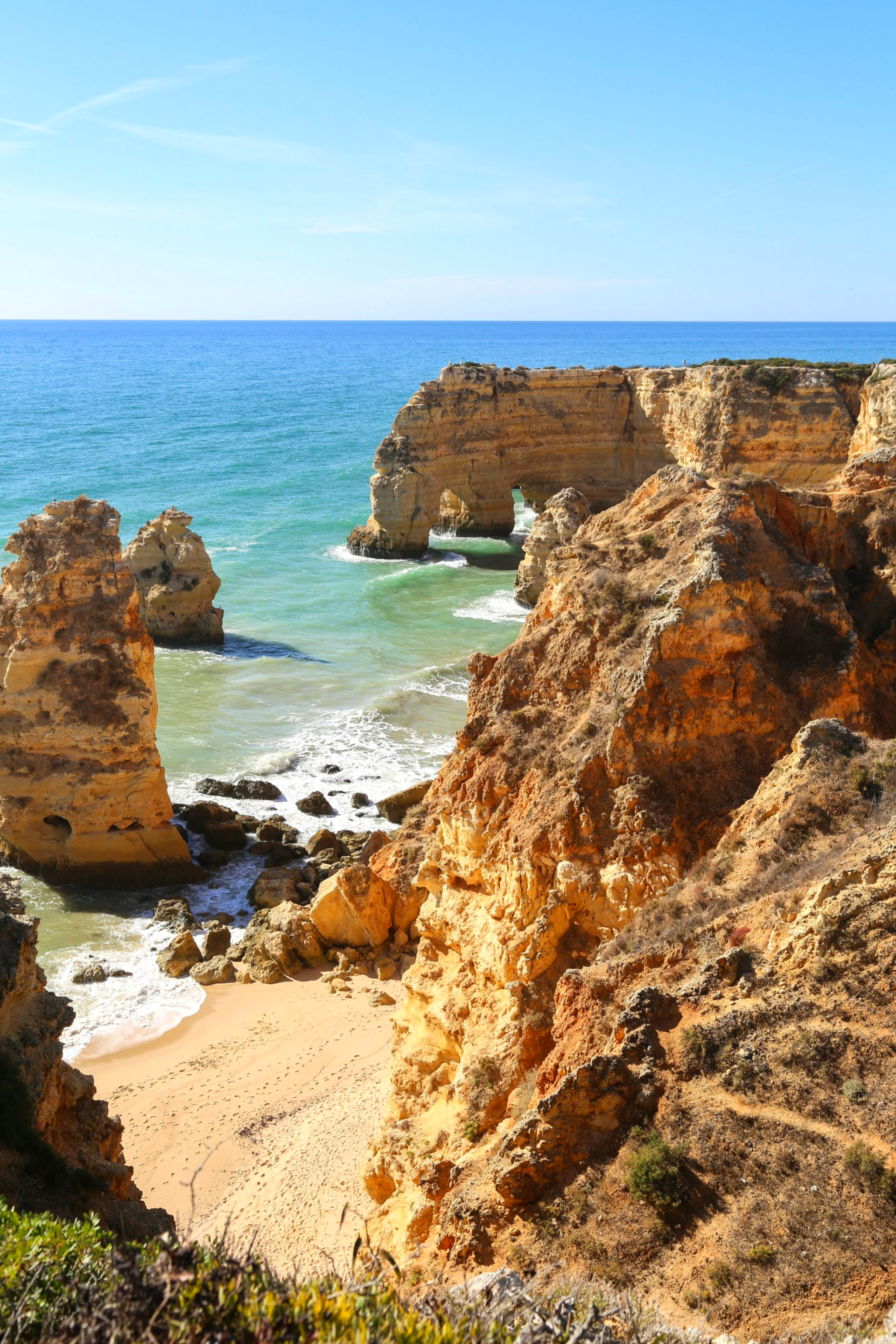 Rocky cliffs with a natural arch overlook a sandy beach and turquoise ocean in The Algarve, Portugal.