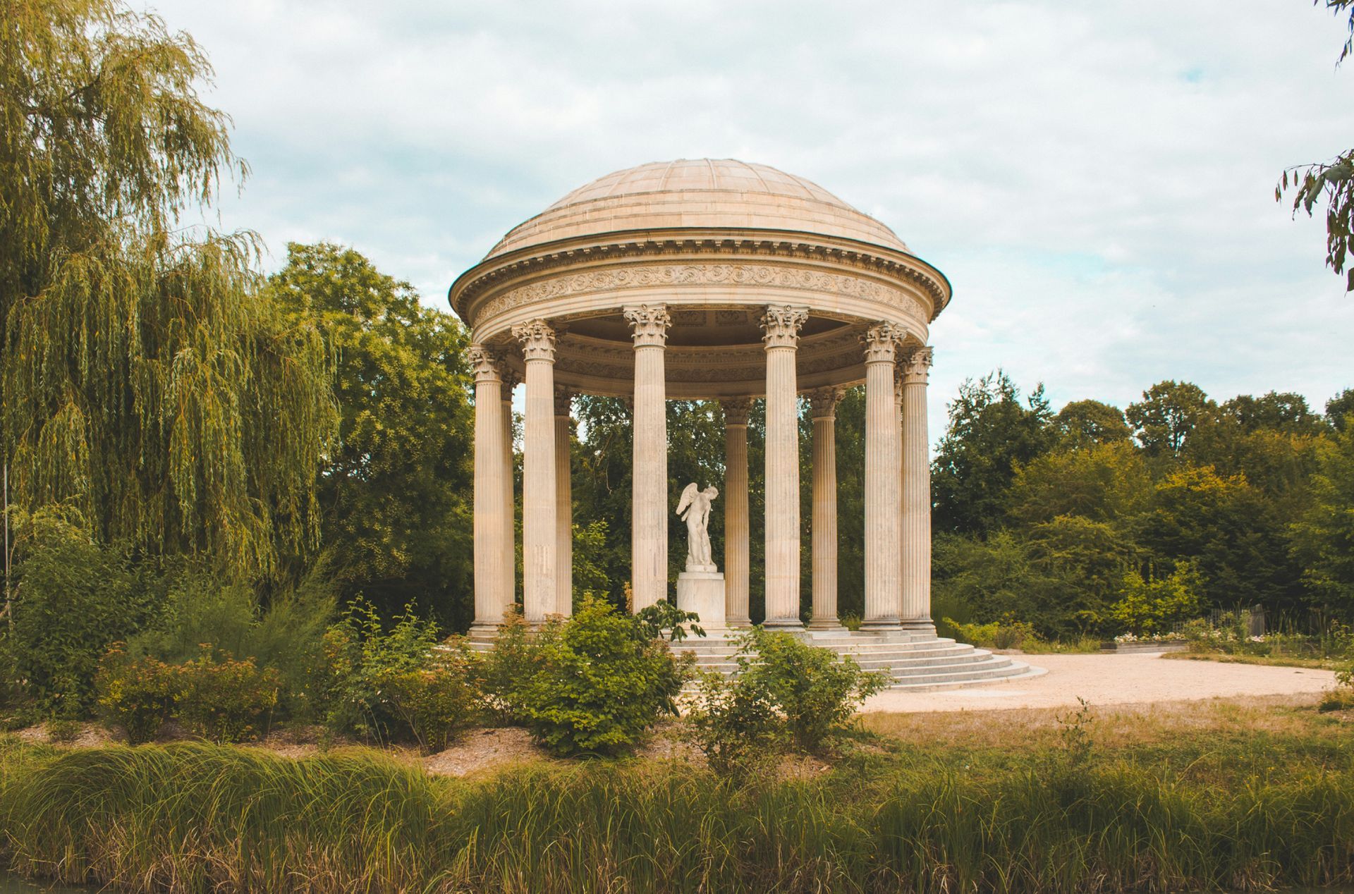 The Temple of Love is a round building with columns and a statue in the middle of a park at Château de Versailles, France.