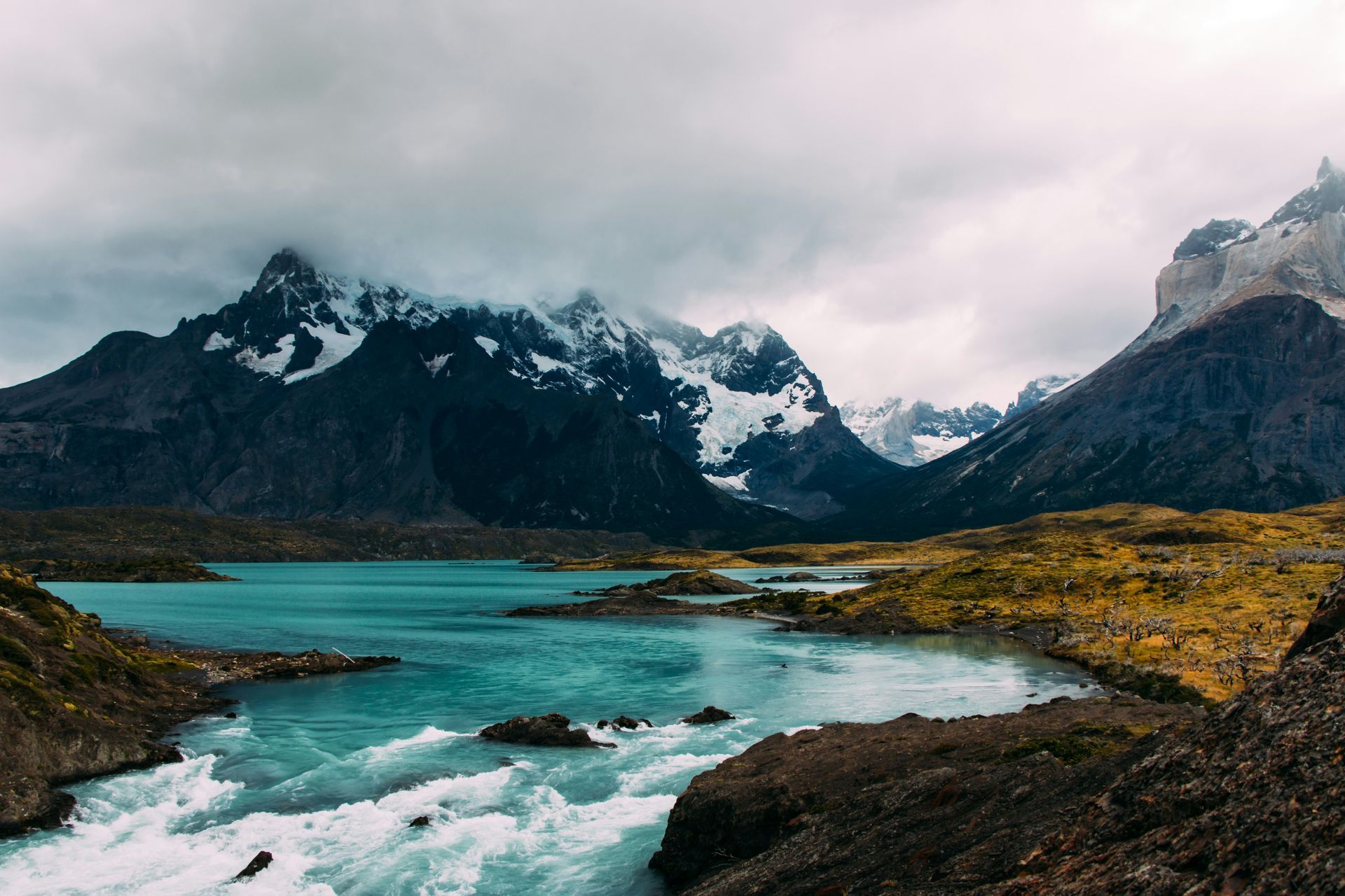Snowy mountains tower over a turquoise lake and rushing river under a cloudy sky in Patagonia, Chile.