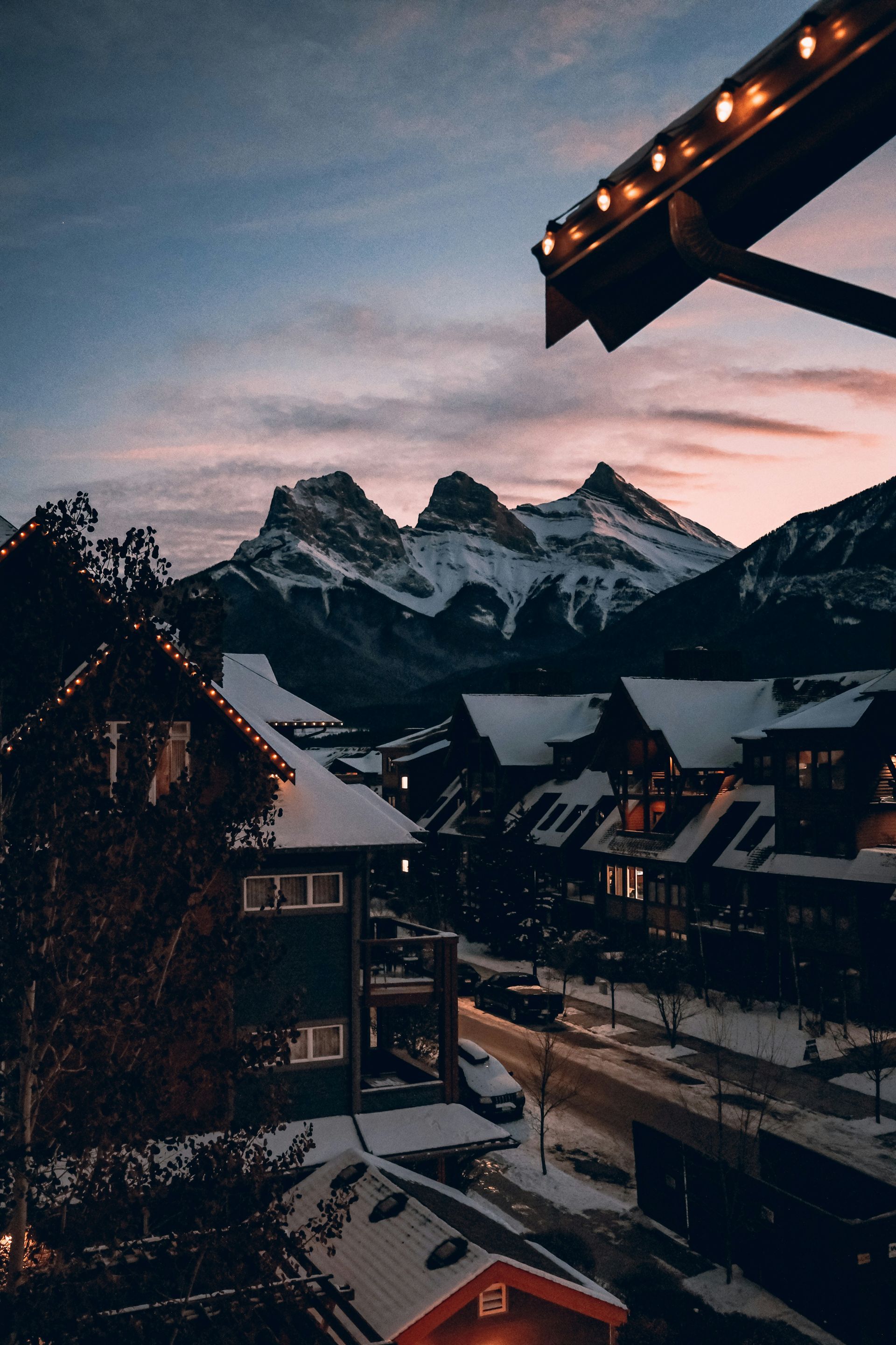 Snow-covered buildings with string lights and street at dusk with mountain range in the background in Canmore, Canada.