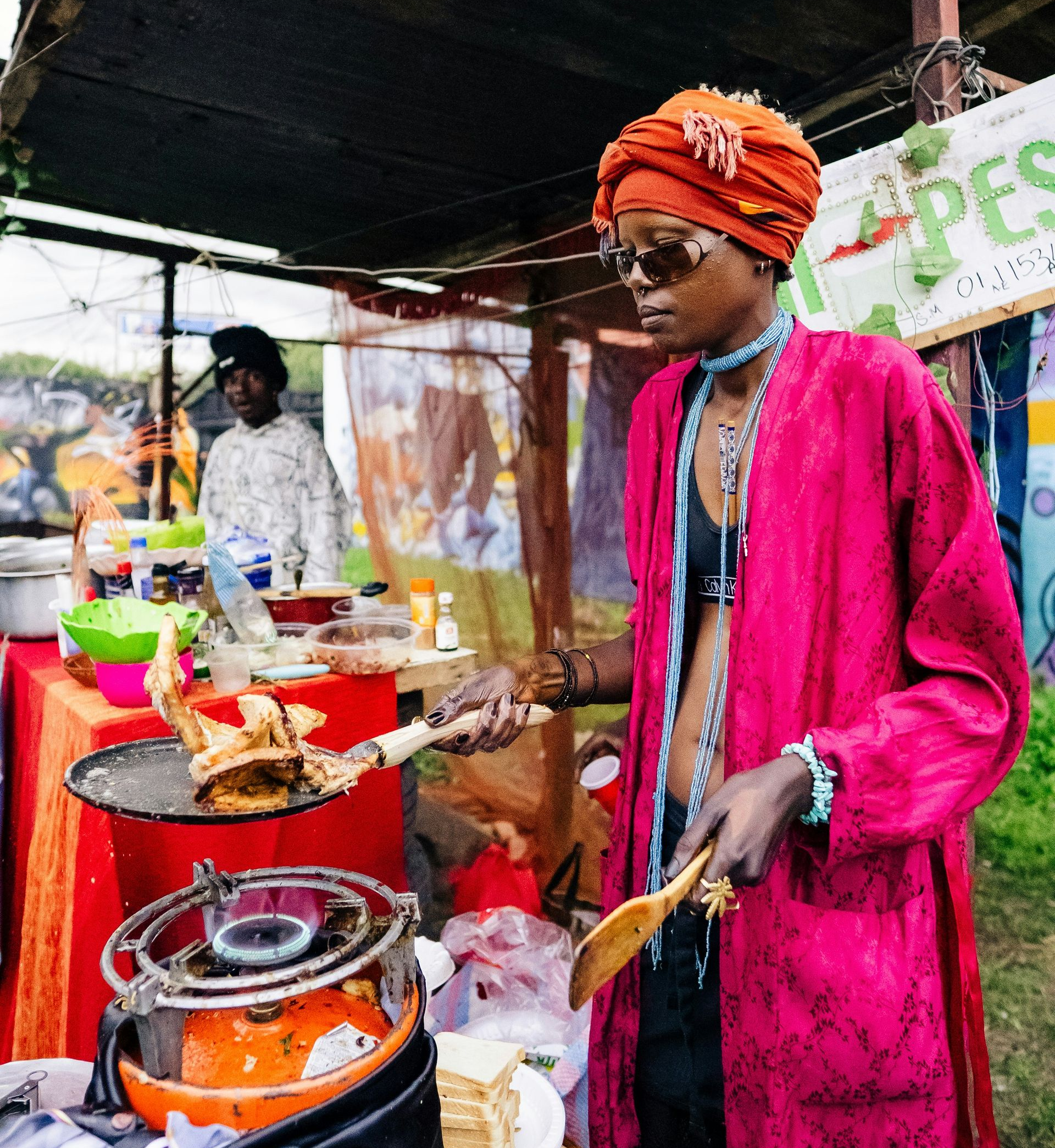 Person in pink robe and turban cooks food at a market stall in Kenya.