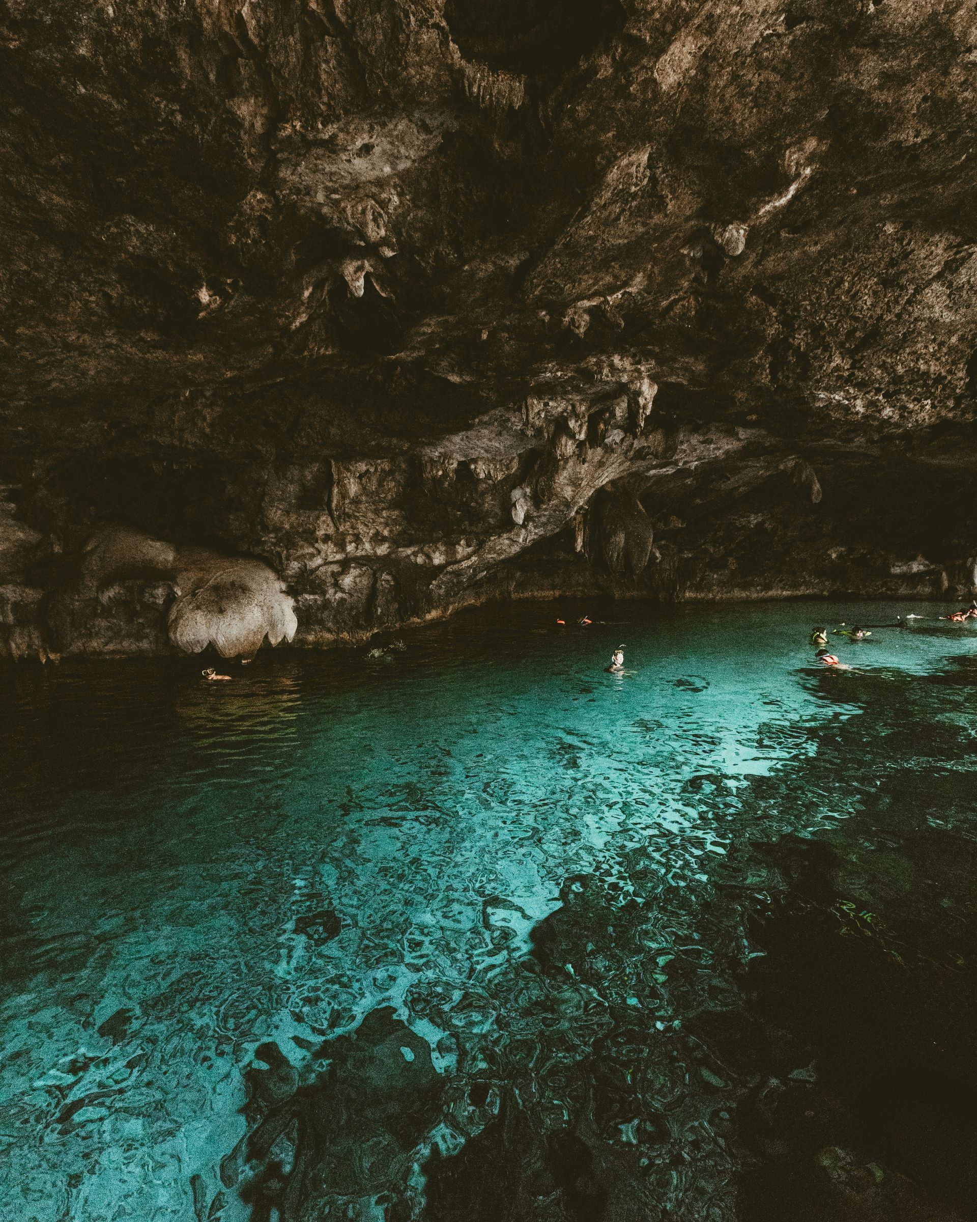 A cenote with turquoise water and swimmers inside a dark, cave-like structure in Tulum, Mexico.