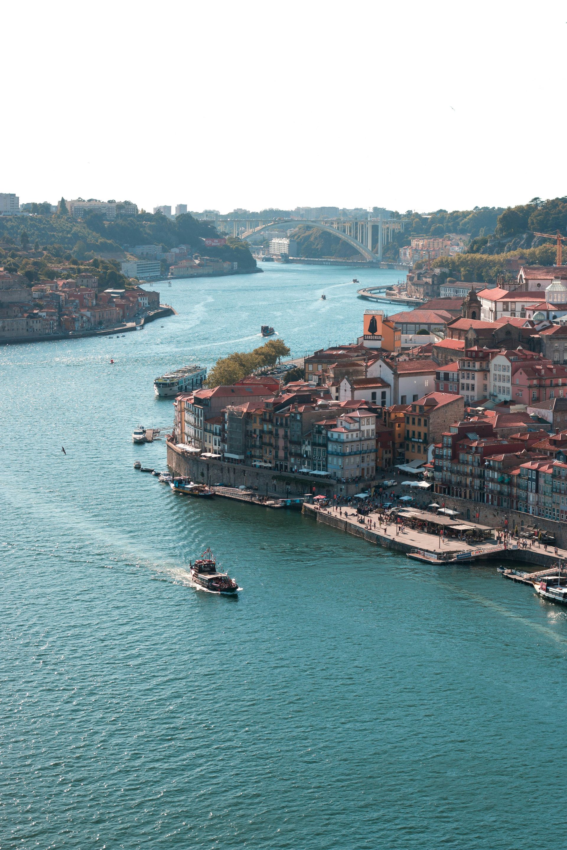 Porto, Portugal: River view with buildings, boats, and a bridge under a bright sky.