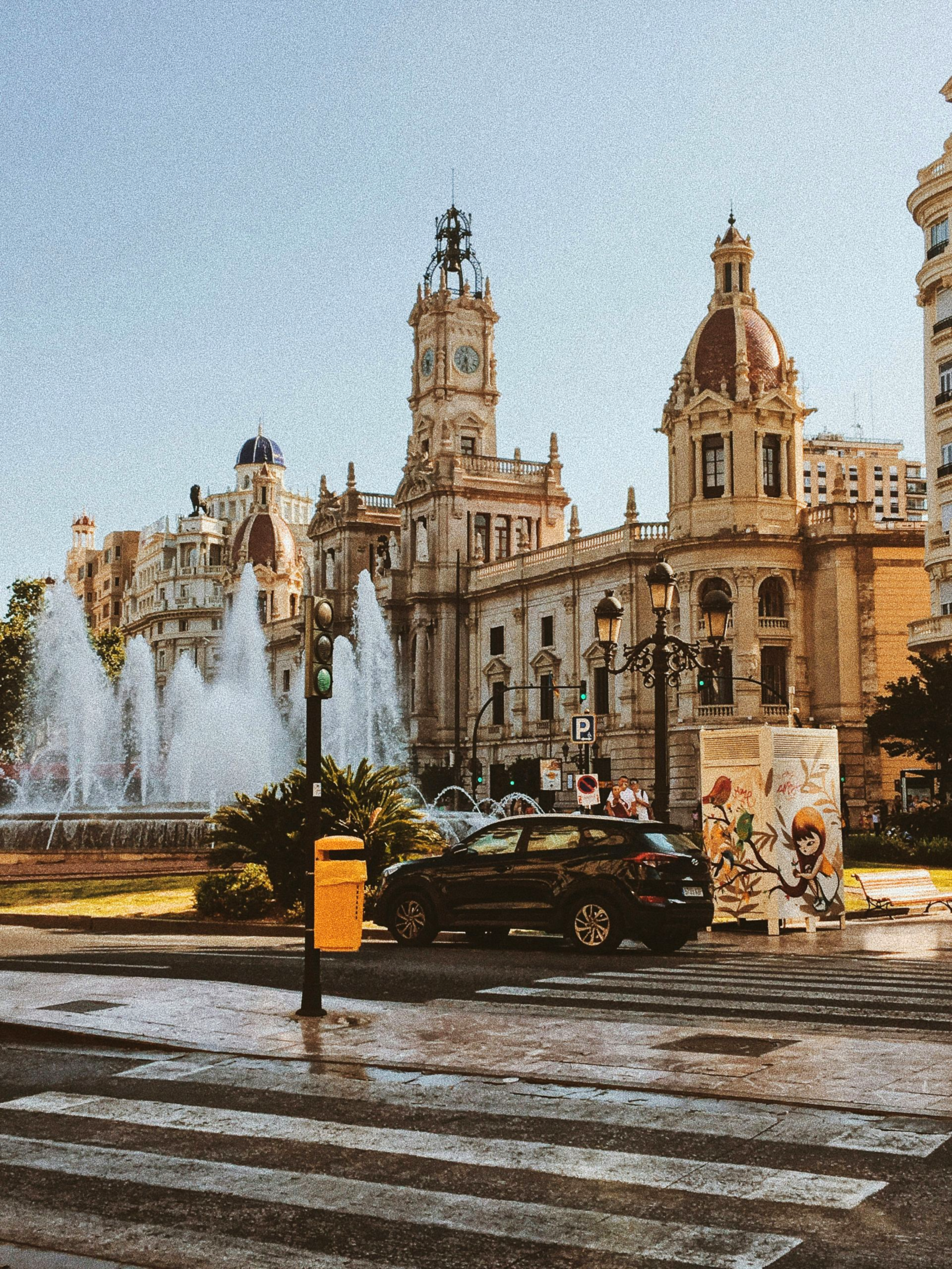 Fountain in a plaza in front of a historic building with a clock tower and car on a wet street in Valencia, Spain.