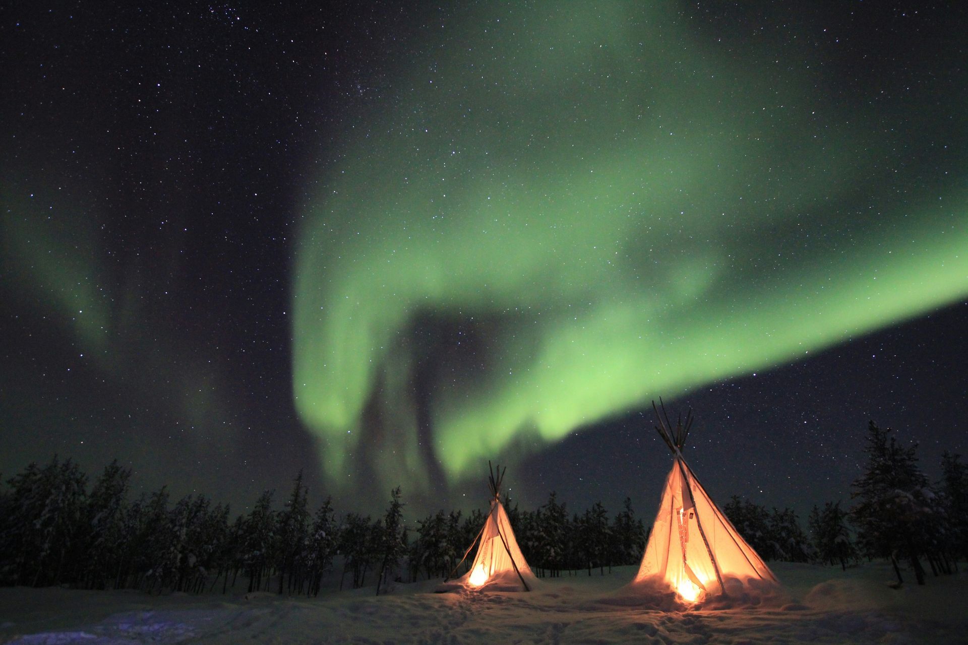 Aurora borealis (Northern Lights) in the night sky above and illuminated teepees in a snowy landscape in Yellowknife, Canada.