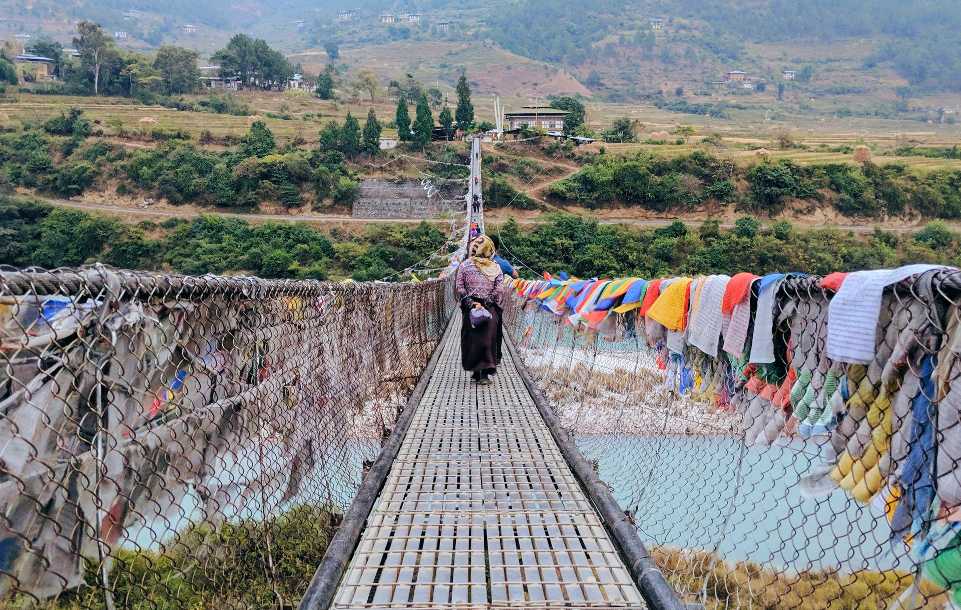 Person walks across a decorated suspension bridge over a turquoise river in Punakha, Bhutan.