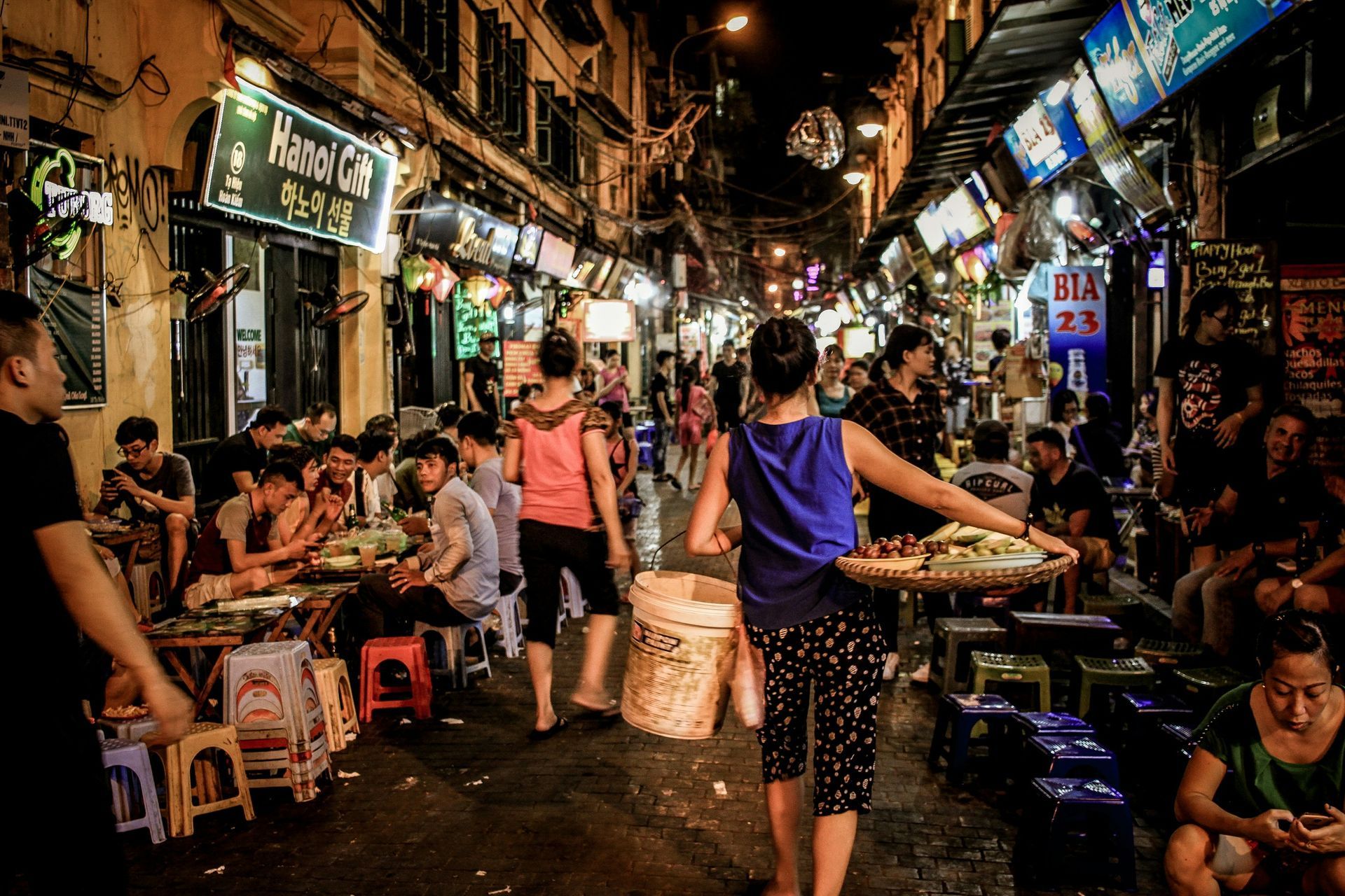 Nighttime crowded alley scene in Hanoi, Vietnam, with people dining and vendors selling food.