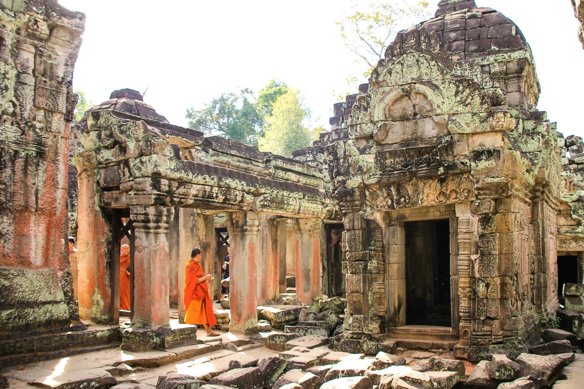 Buddhist monks in orange robes walk through ancient stone temple ruins in sunlight in Siem Reap, Cambodia.