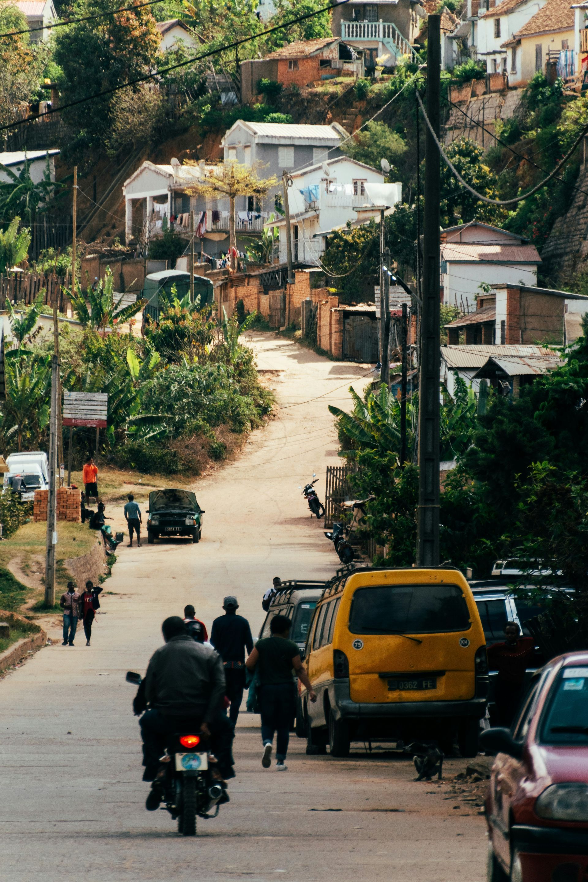 A bustling street in Ambositra, Madagascar, with people walking and vehicles driving. Yellow van and motorcycles in focus.