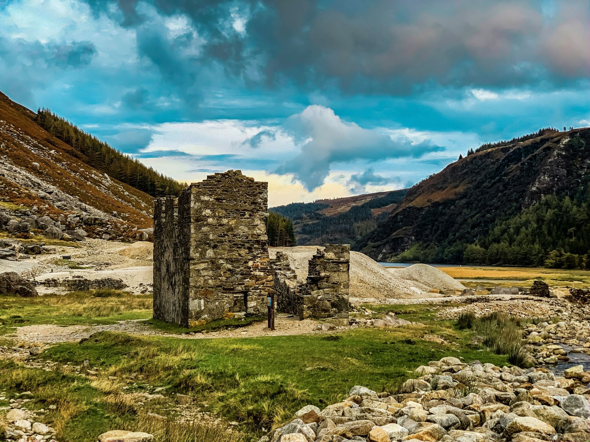 Ruined stone building in a valley with mountains, green grass, rocks, and cloudy blue sky in Glendalough, Ireland.