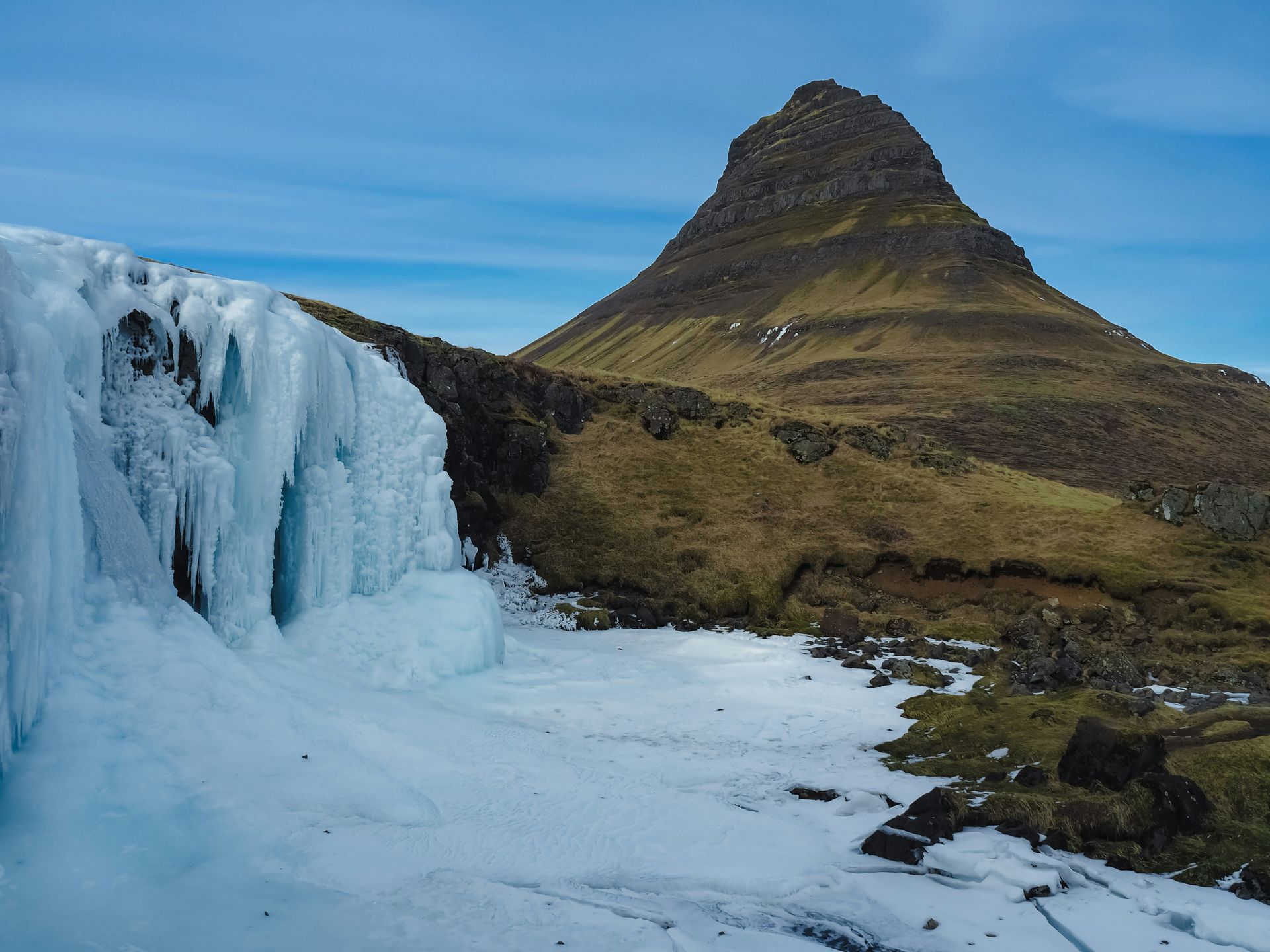 Frozen waterfall in Iceland, with the conical Kirkjufell mountain in the background under a blue sky in Snæfellsnes, Iceland.