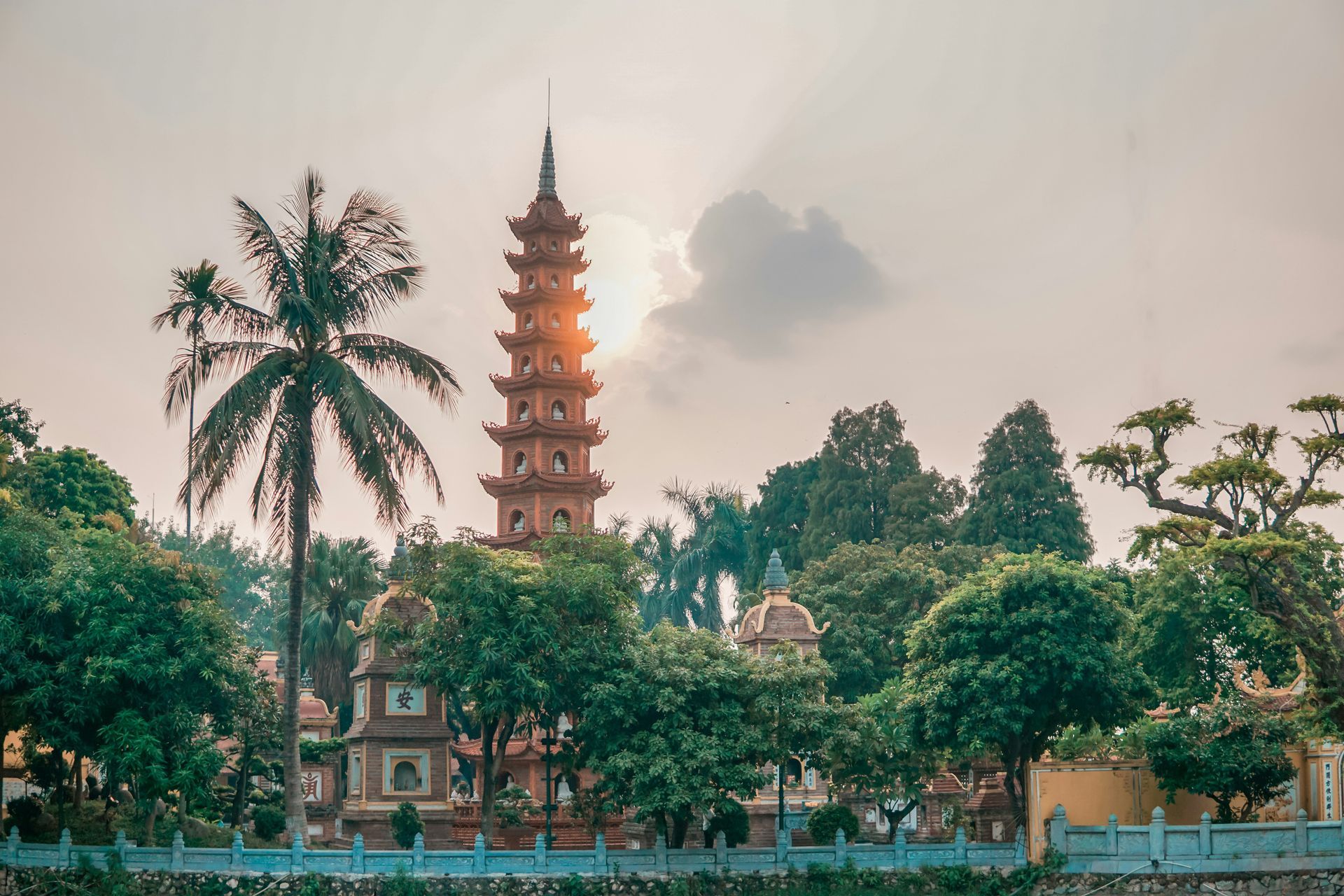 A tall pagoda with the sun shining behind it, surrounded by trees, in Hanoi, Vietnam.