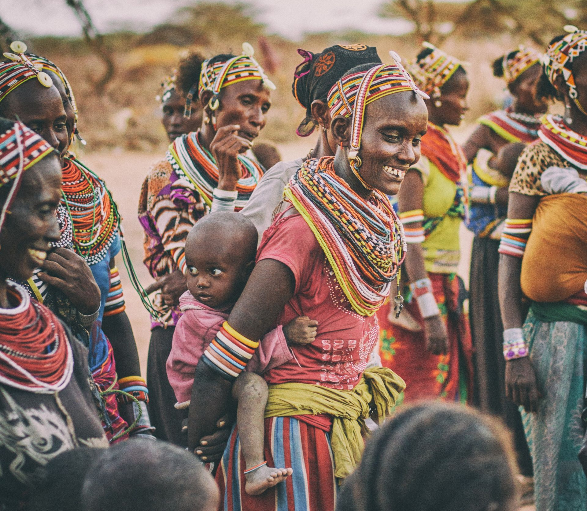 Group of smiling nomadic women, wearing traditional, colorful beaded jewelry and carrying a child outdoors in Kargi, Kenya.