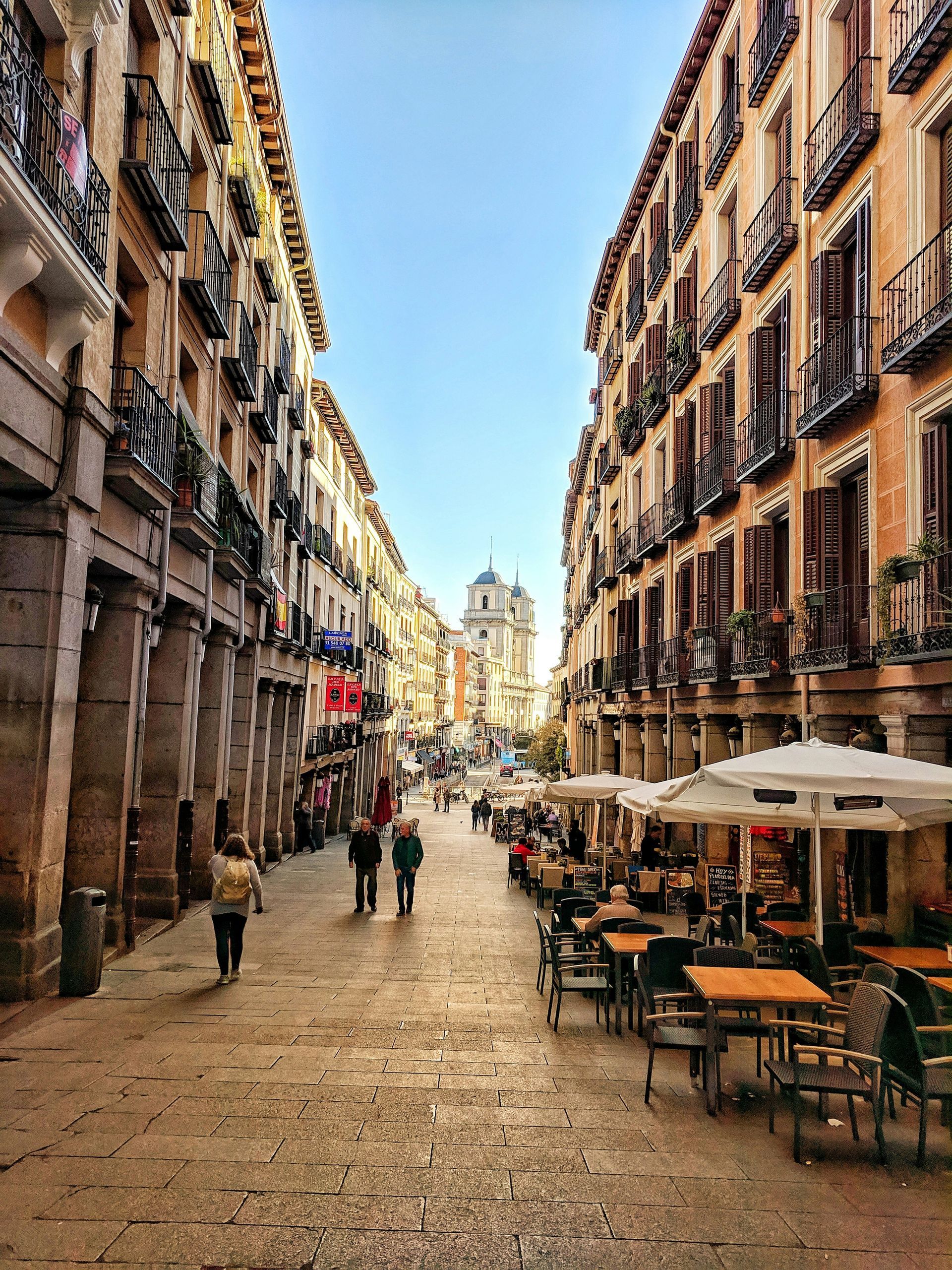 Cobblestone street in Madrid, Spain, lined with restaurants and historic buildings. People walk and sit at tables.
