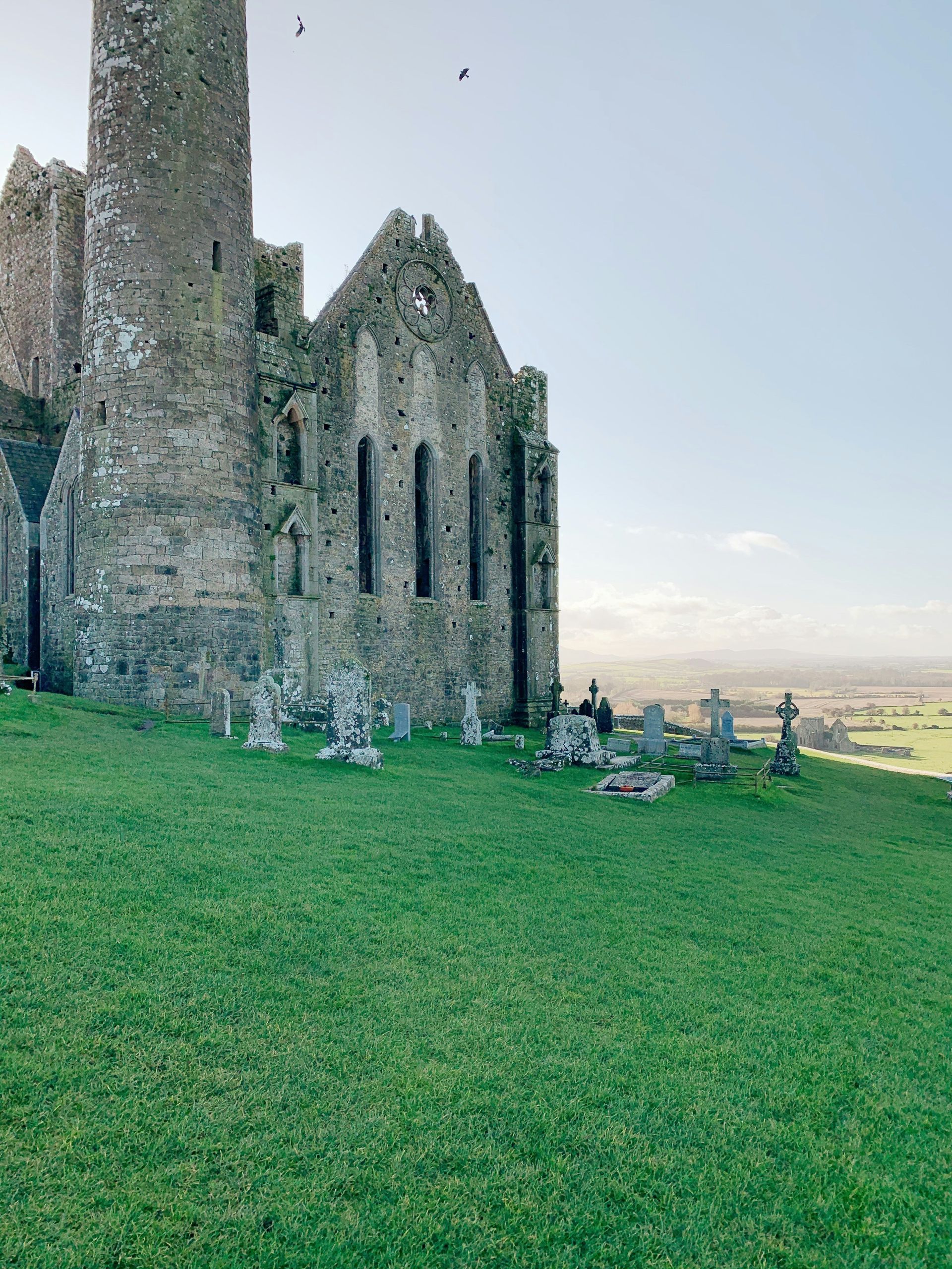 Ancient stone ruins on a green hillside, overlooking a distant landscape under a blue sky at the Rock of Cashel, Ireland.