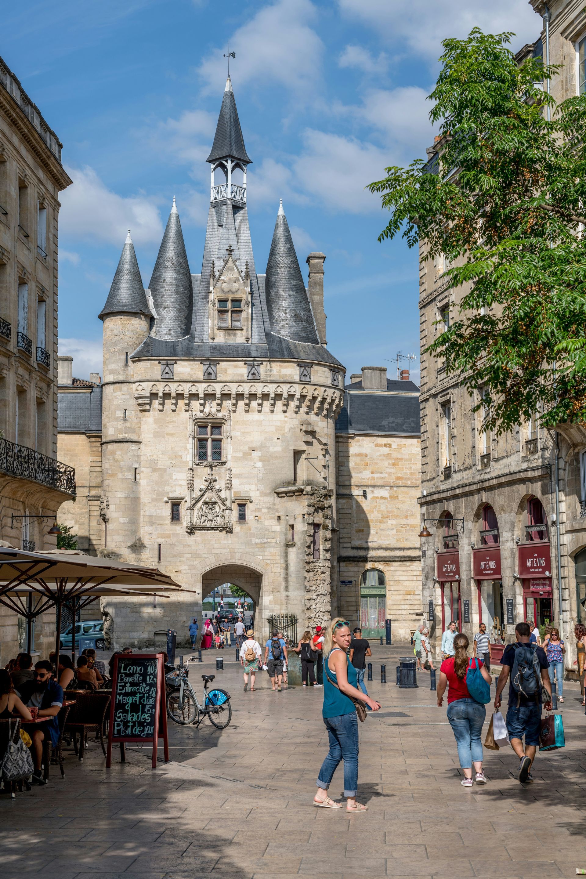 A group of people are walking down a street in front of the Porte Cailhau in Bordeaux, France.