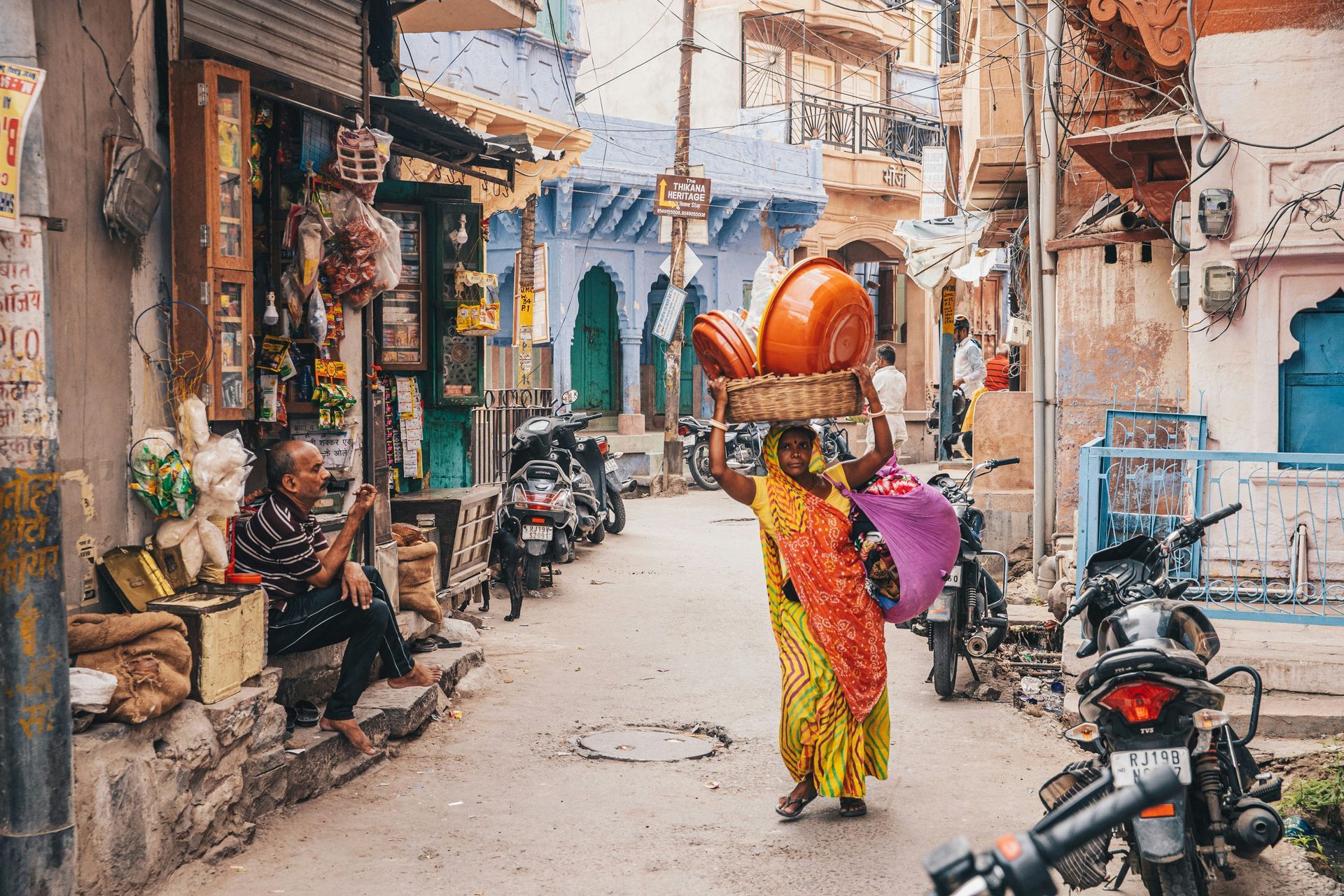 Woman carries bowls on her head through a bustling street in India, lined with shops, motorcycles, and colorful buildings.