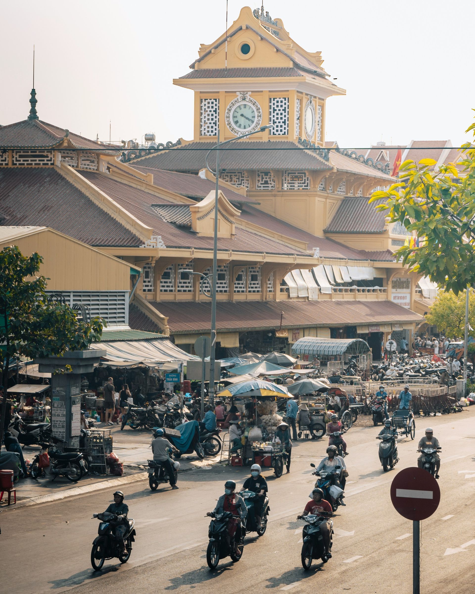 Yellow-colored market building with clock tower, busy street scene in front, many motorcycles in Ho Chi Minh, Vietnam.