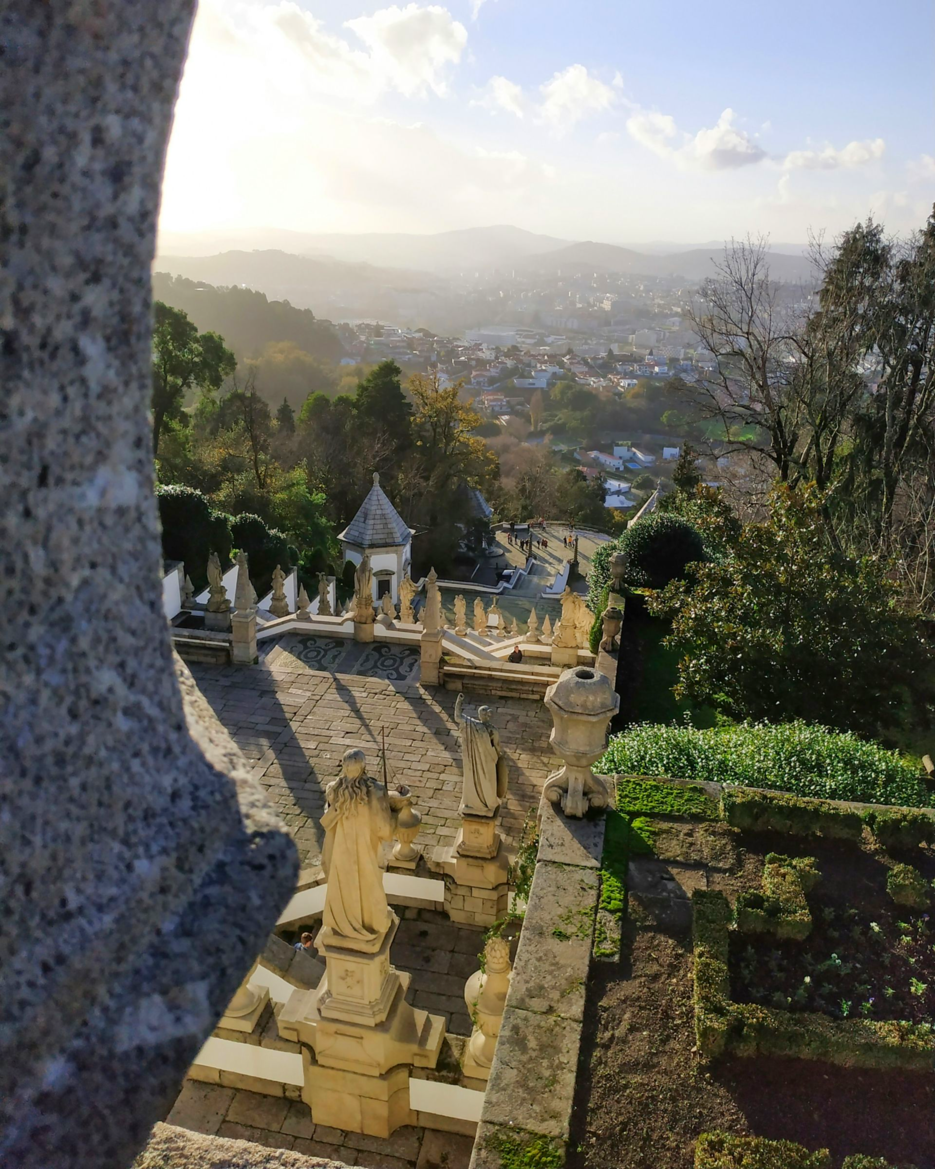 Stone staircase with statues overlooks a valley town, trees and hazy mountains under a sunny sky in Braga, Portugal.