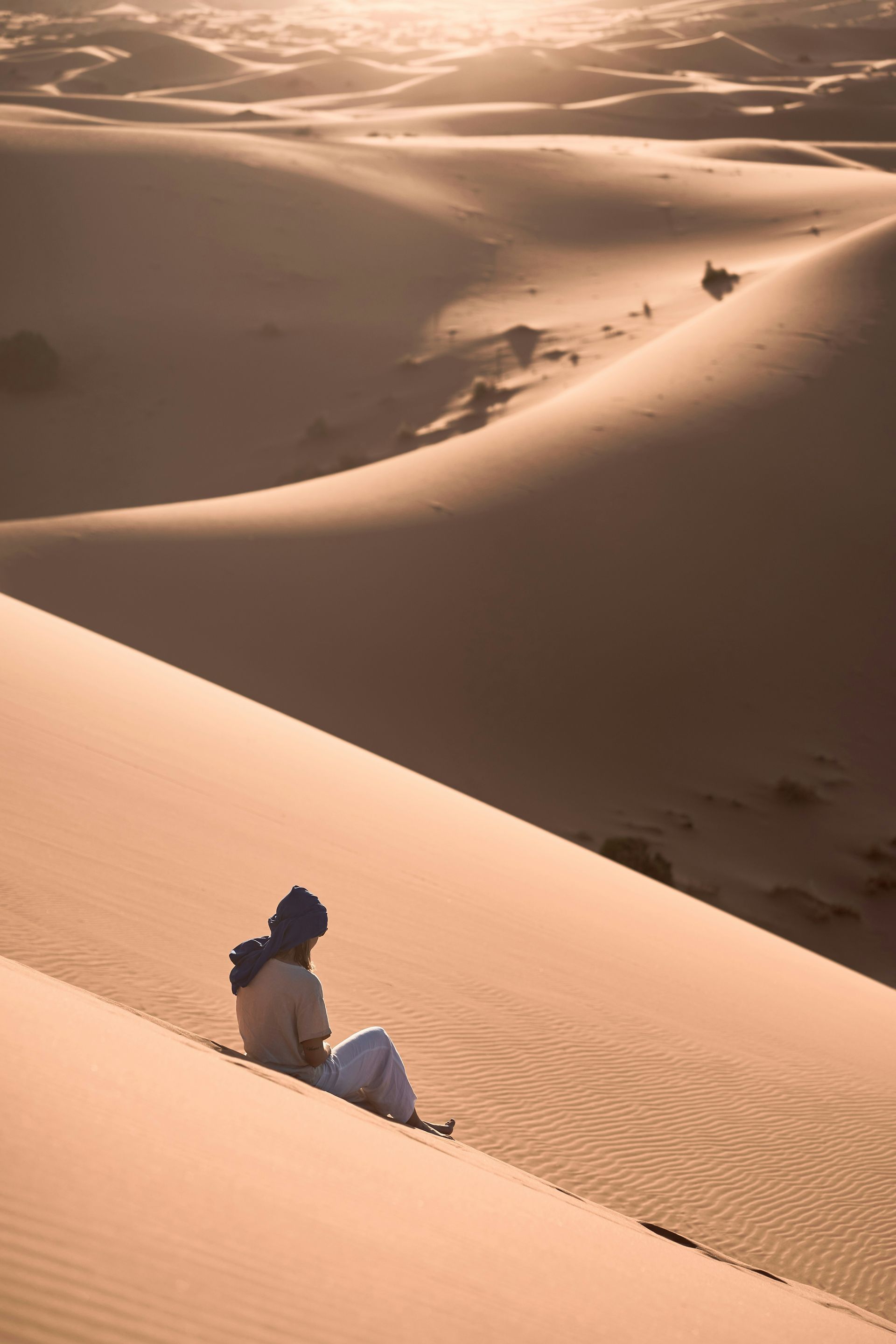 A person is sitting on top of a sand dune in the desert in Erg Chebbi, Morocco.